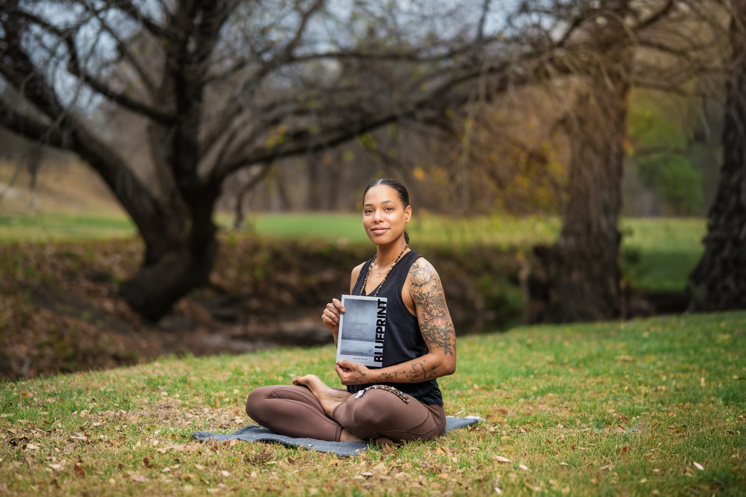 Woman sitting on grass holding a book outdoors with trees in background.