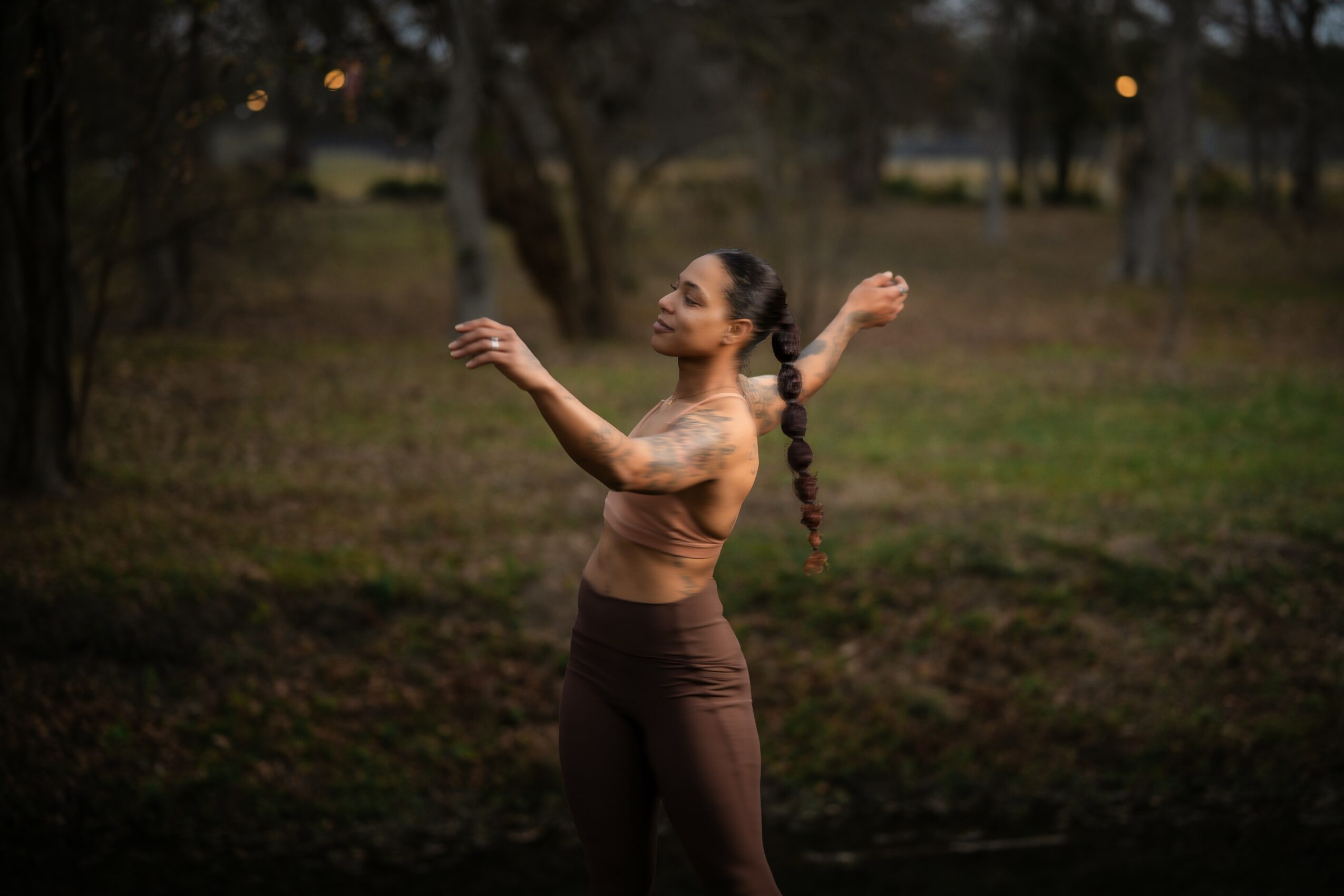 Woman with long braided hair standing outdoors with arms raised, in a natural setting with trees and grass.