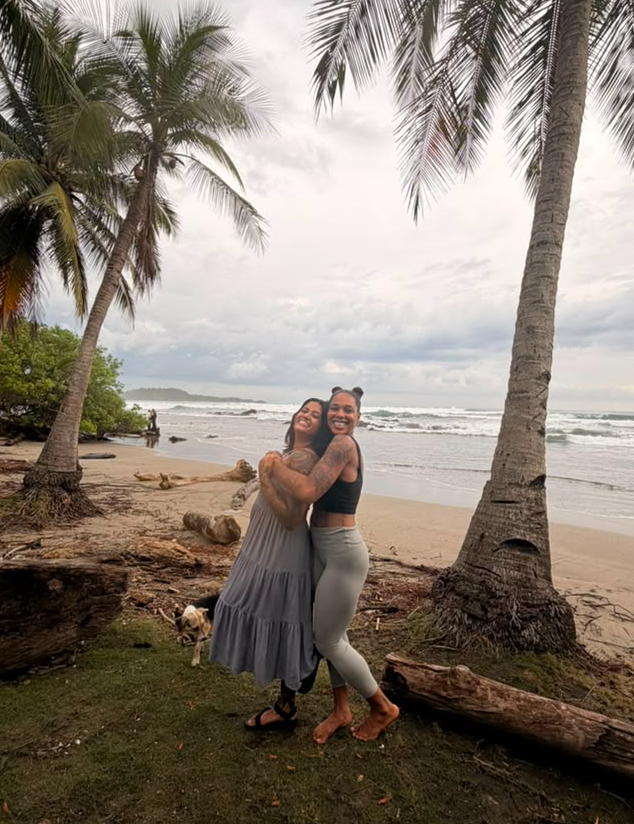 Two women hugging on a beach with palm trees and ocean in the background.