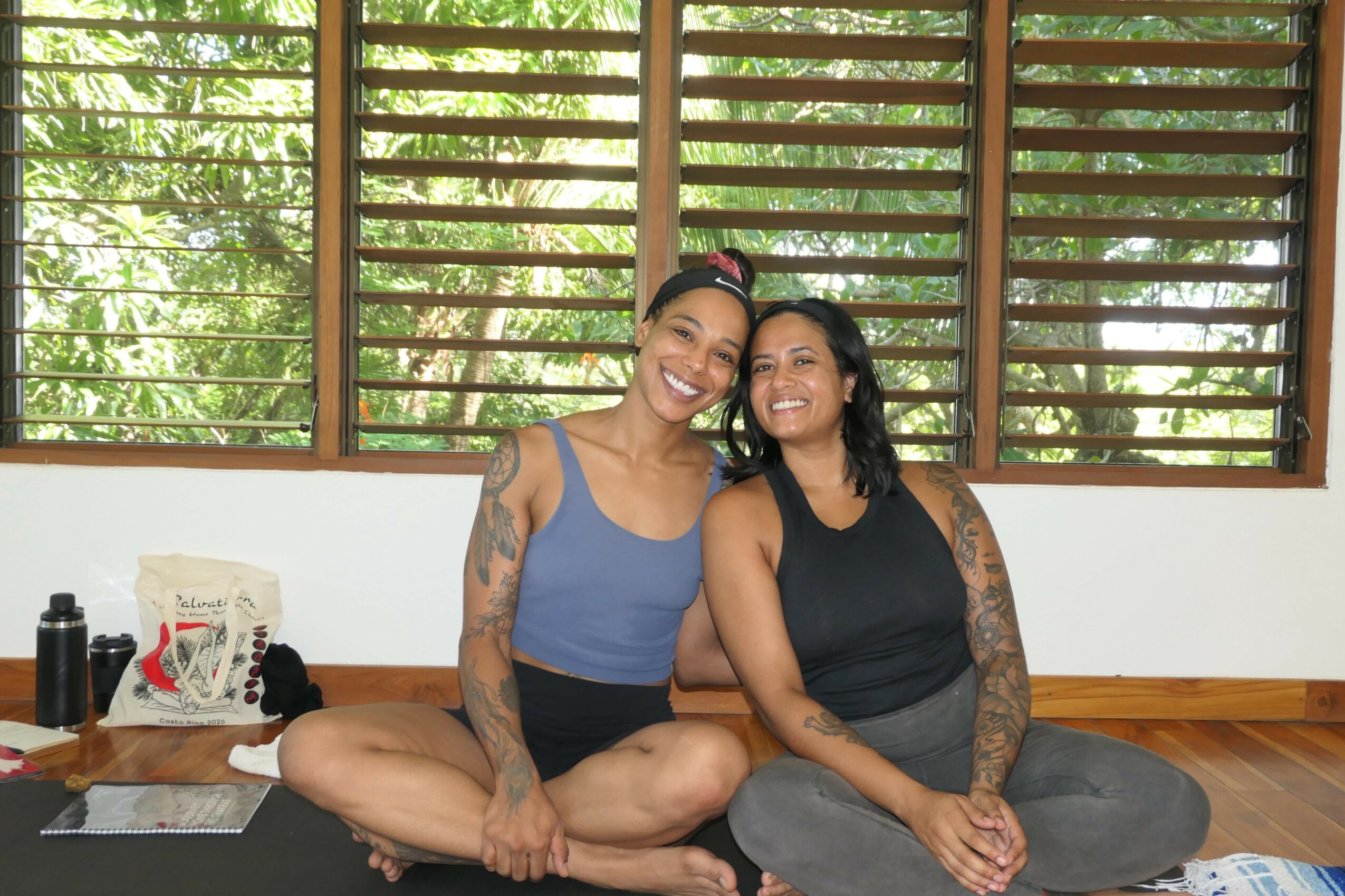 Two women sit on a wooden floor in front of a window with wooden blinds, smiling and leaning toward each other.