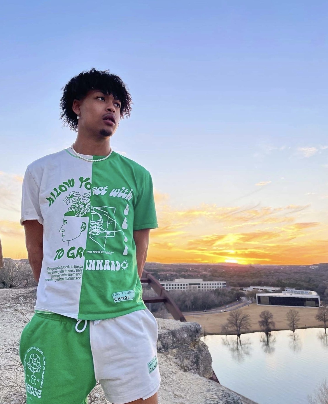 Young man with curly hair wearing a green and white t-shirt and white shorts, standing outdoors near a body of water at sunset.