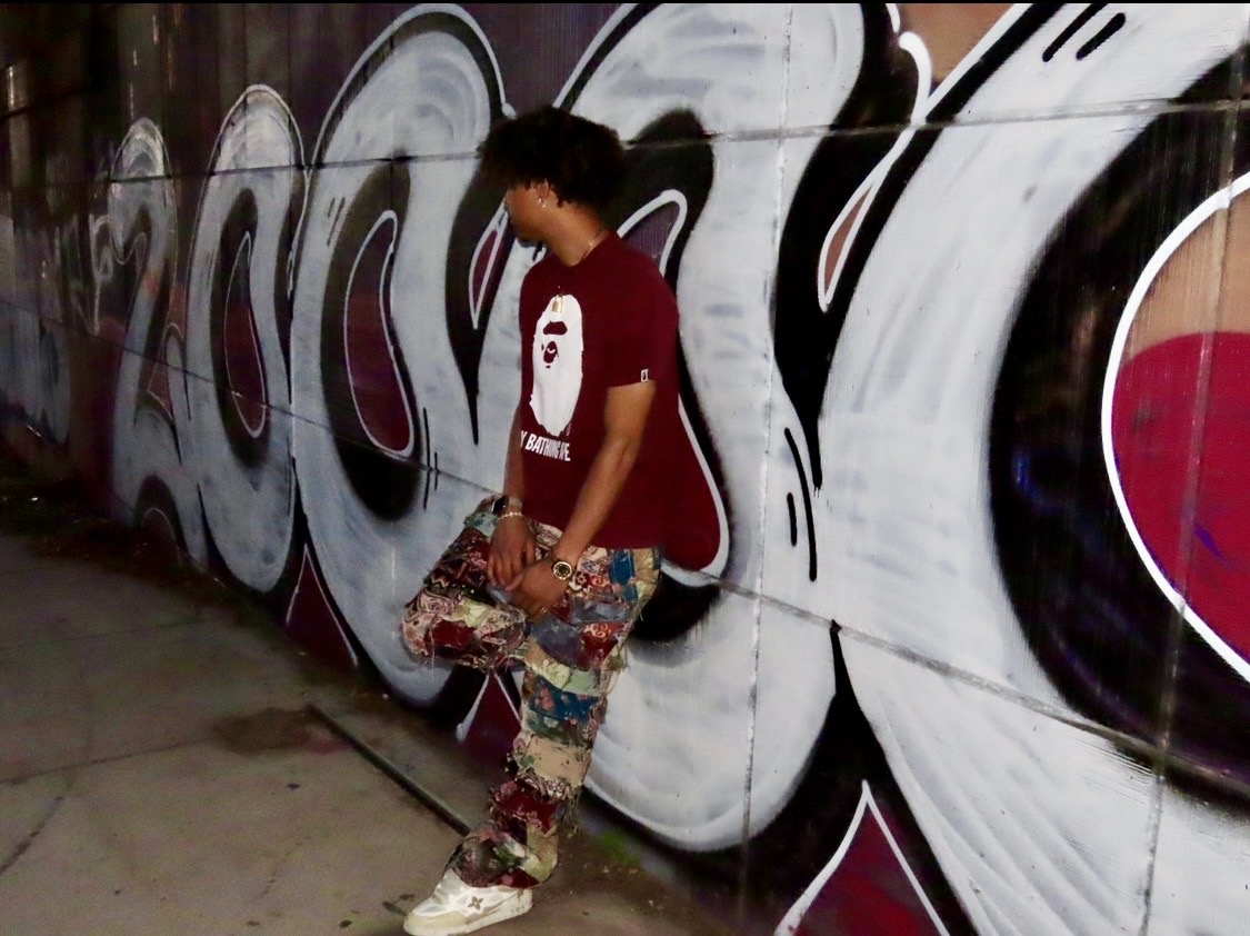 Person standing against graffiti wall holding a skateboard, wearing a red shirt and patterned pants.