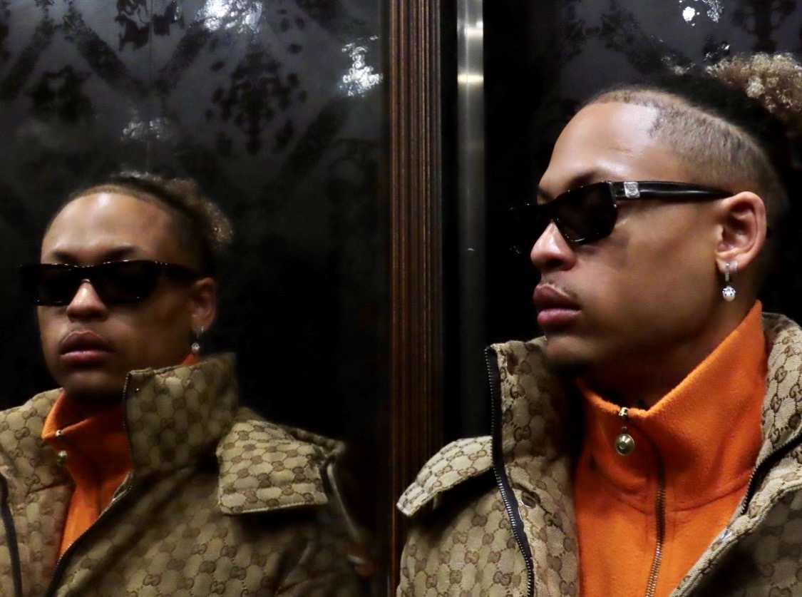 Two men wearing sunglasses and patterned jackets, standing in front of a reflective surface, with one facing slightly to the right.