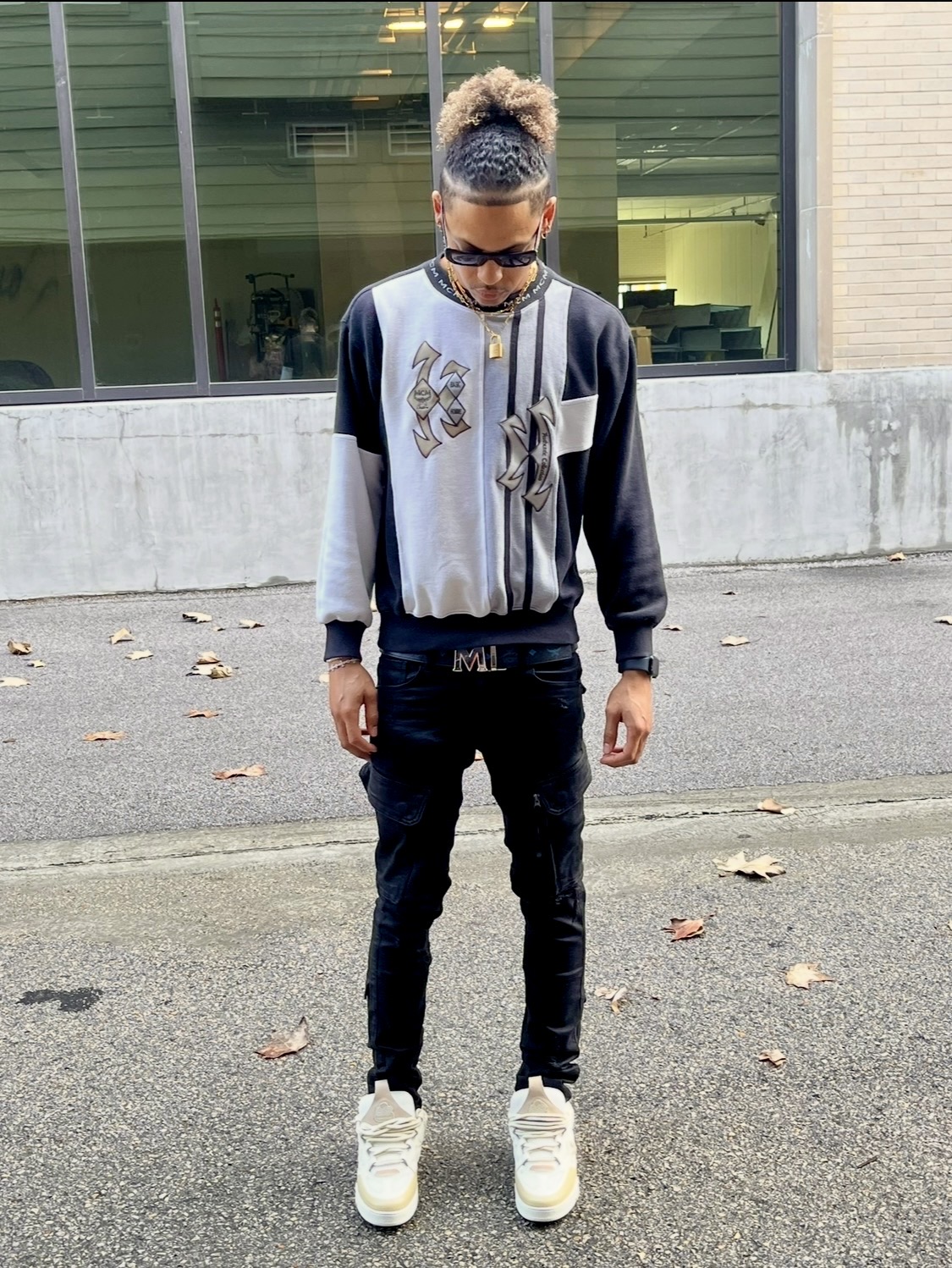 Young person standing outdoors on pavement with fallen leaves, wearing glasses, a color-blocked jacket, black pants, and white sneakers.