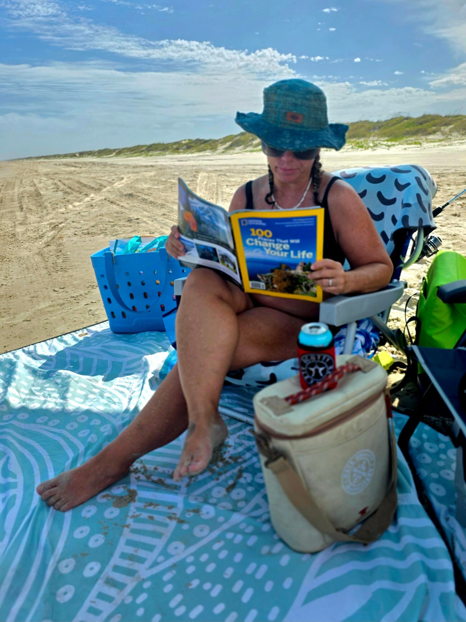 Child sitting on a beach towel reading a book, wearing a large sun hat, with a bag and water bottle nearby, sandy beach and sky in background.