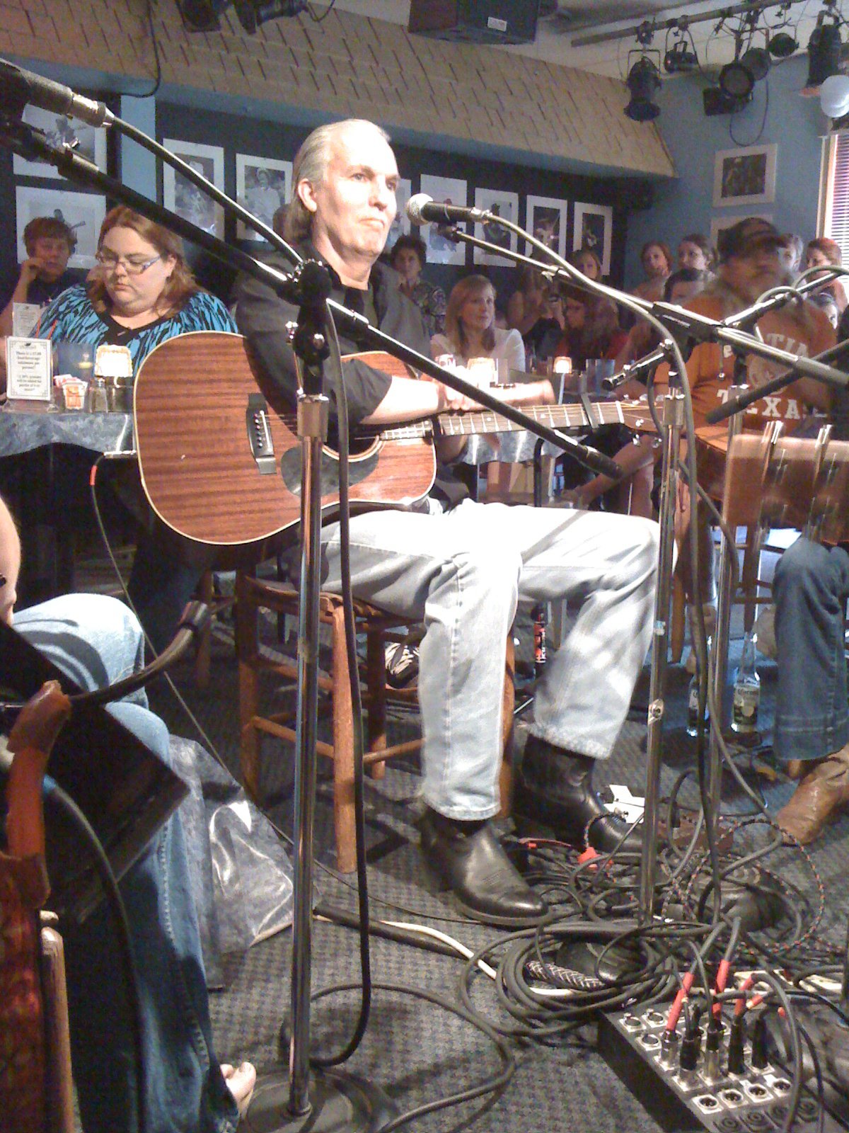 Man sitting with guitar, surrounded by microphones, in a lively indoor setting with people and framed pictures on the wall.