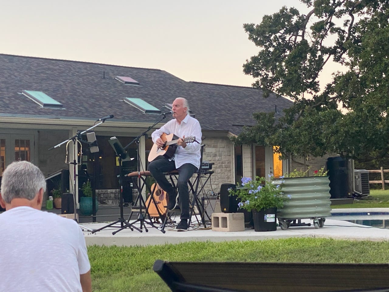 Person playing guitar on an outdoor stage with a house and trees in the background.
