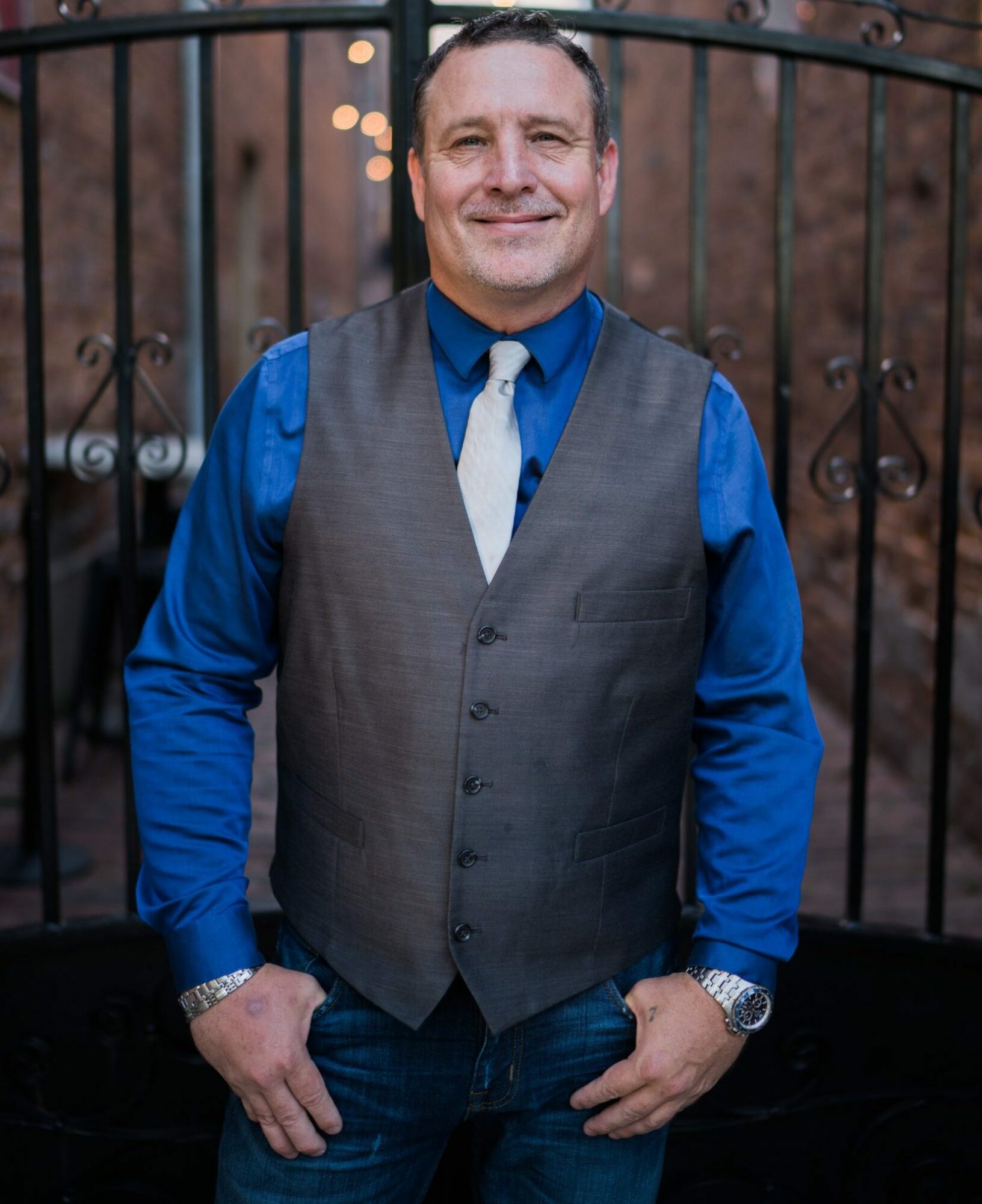 Man in blue shirt and gray vest standing outdoors with a black fence behind him, smiling, with blurred trees in background.