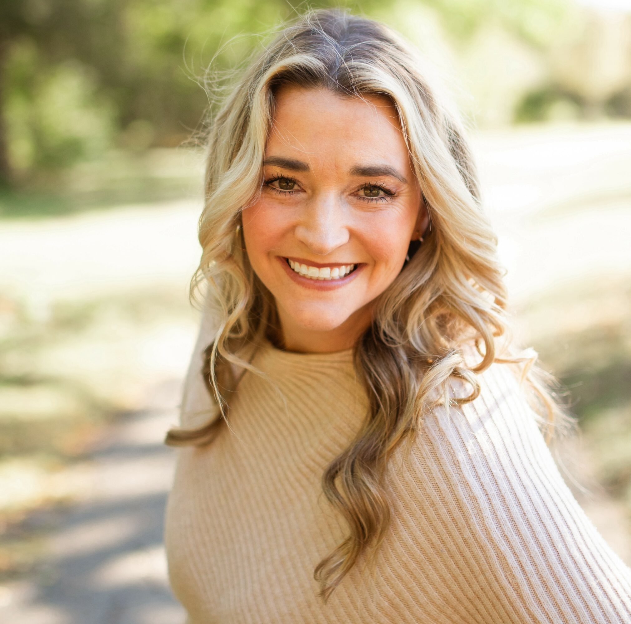 Smiling woman with wavy blonde hair outdoors, wearing a beige sweater, sunny background with trees.