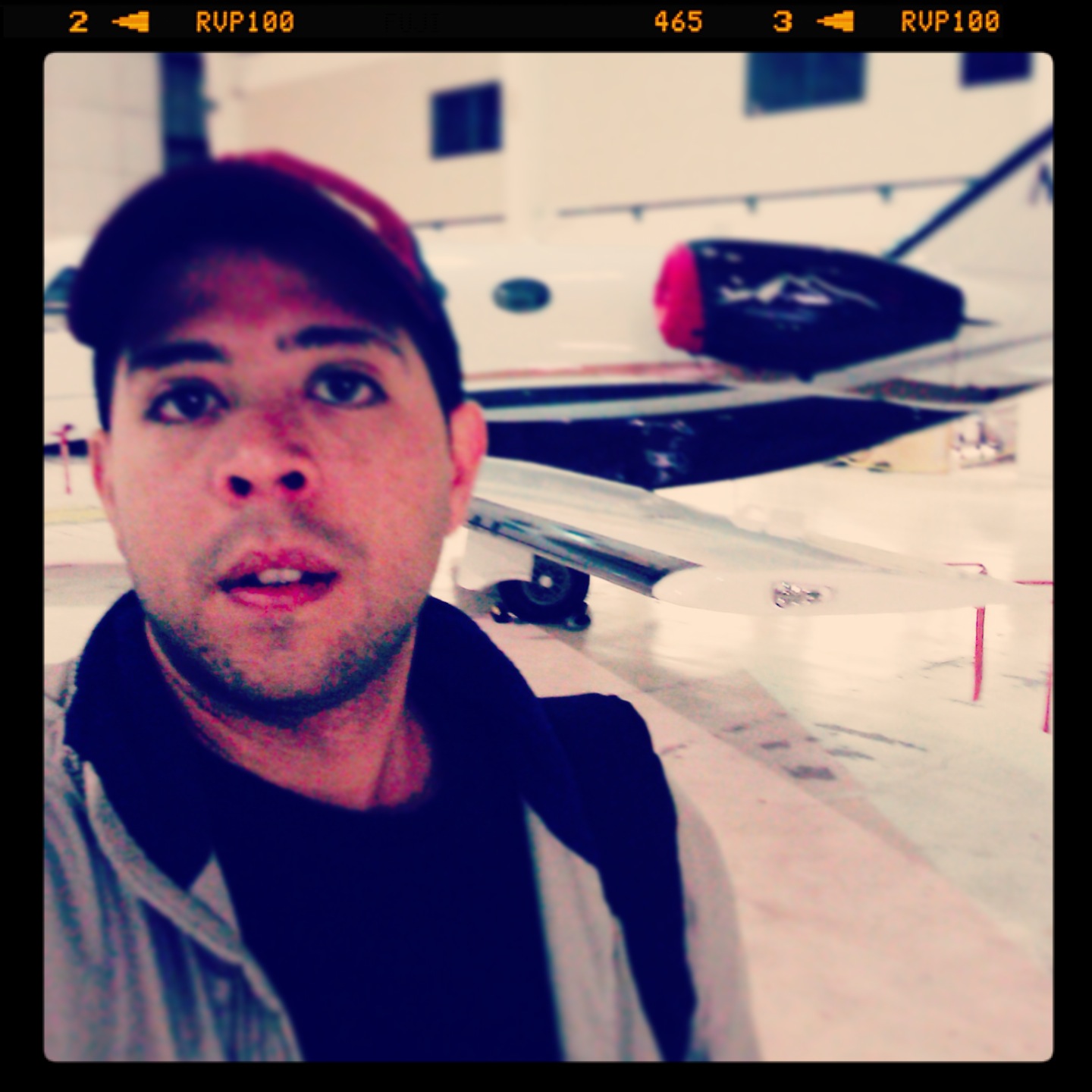 Person taking selfie outdoors with a boat and building in background, wearing a cap and jacket.