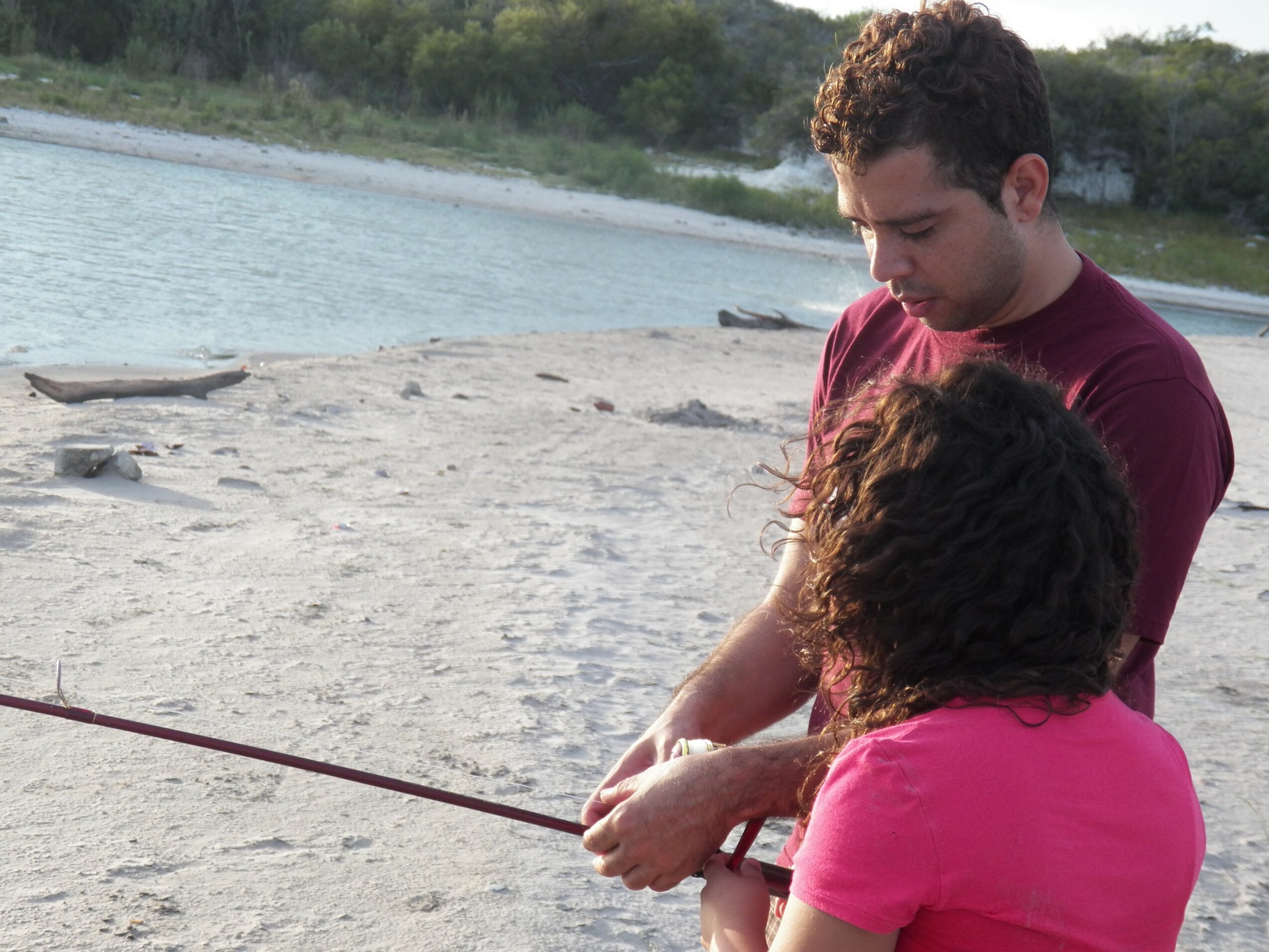 Two people, a man and a woman, are on a sandy beach near water, holding a fishing rod together.
