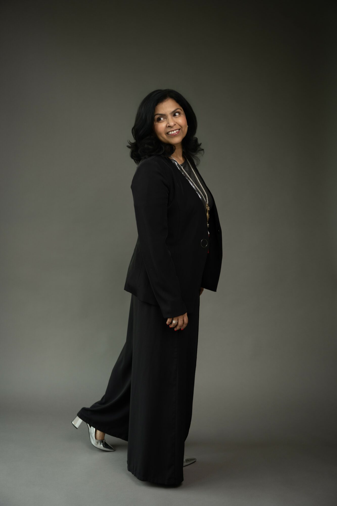 Woman in black suit standing against gray background, smiling, with shoulder-length dark hair, wearing jewelry.