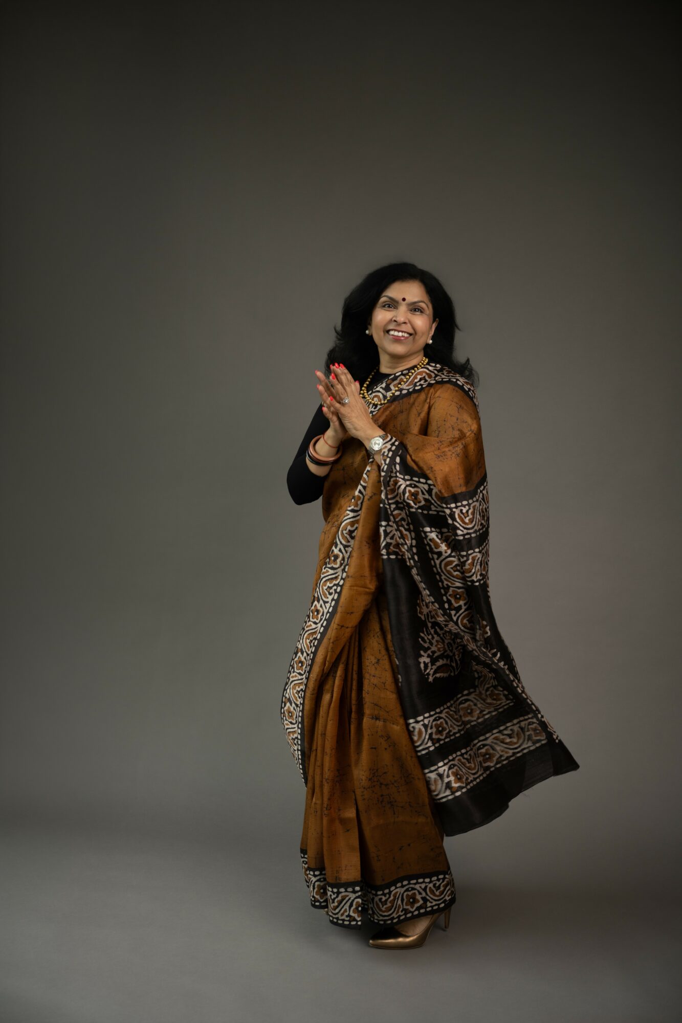 Woman in traditional attire smiling with hands together, standing against a plain background.