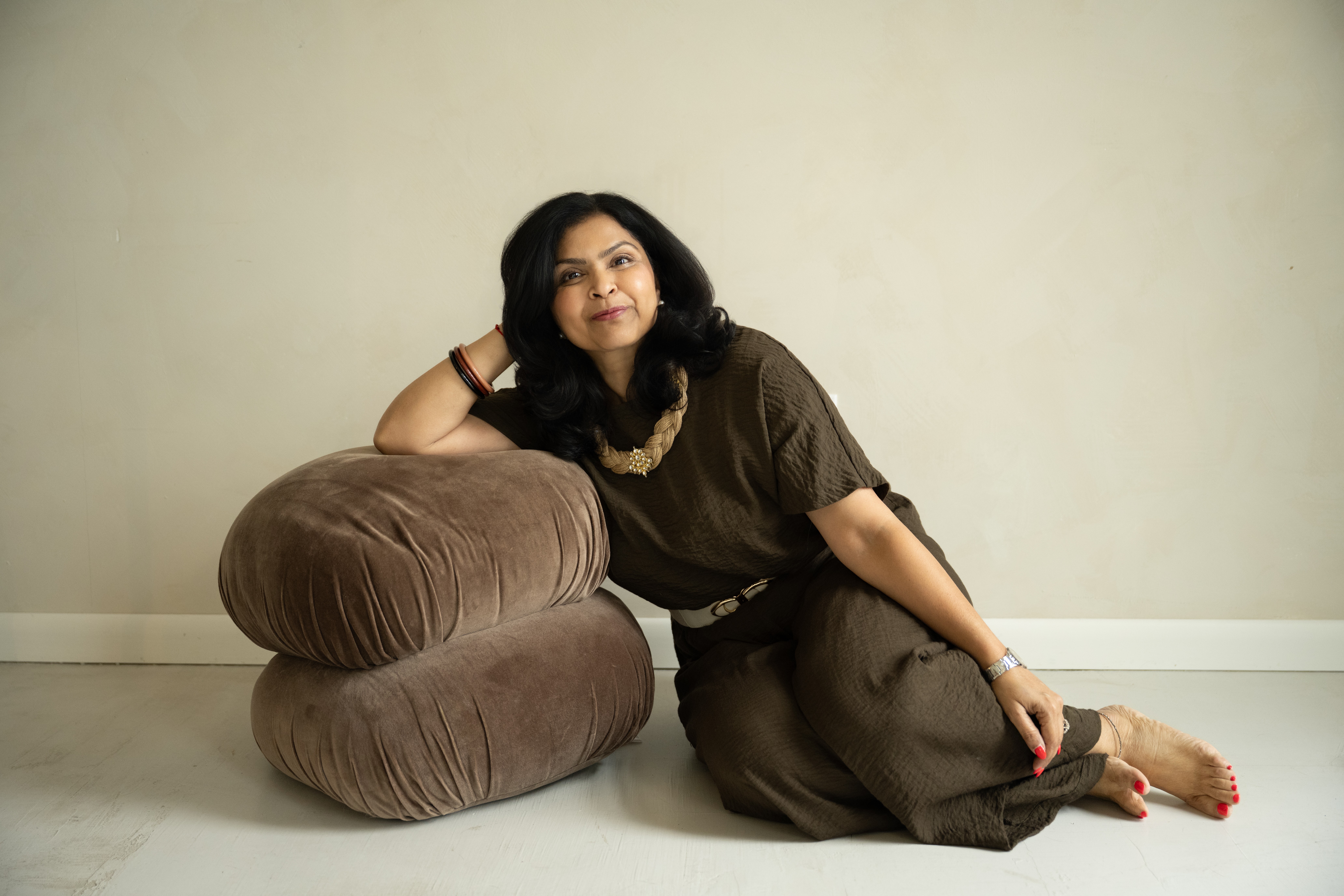 Woman with dark hair lying on the floor, resting her head on a large brown cushion, wearing a dark outfit, against a plain wall.