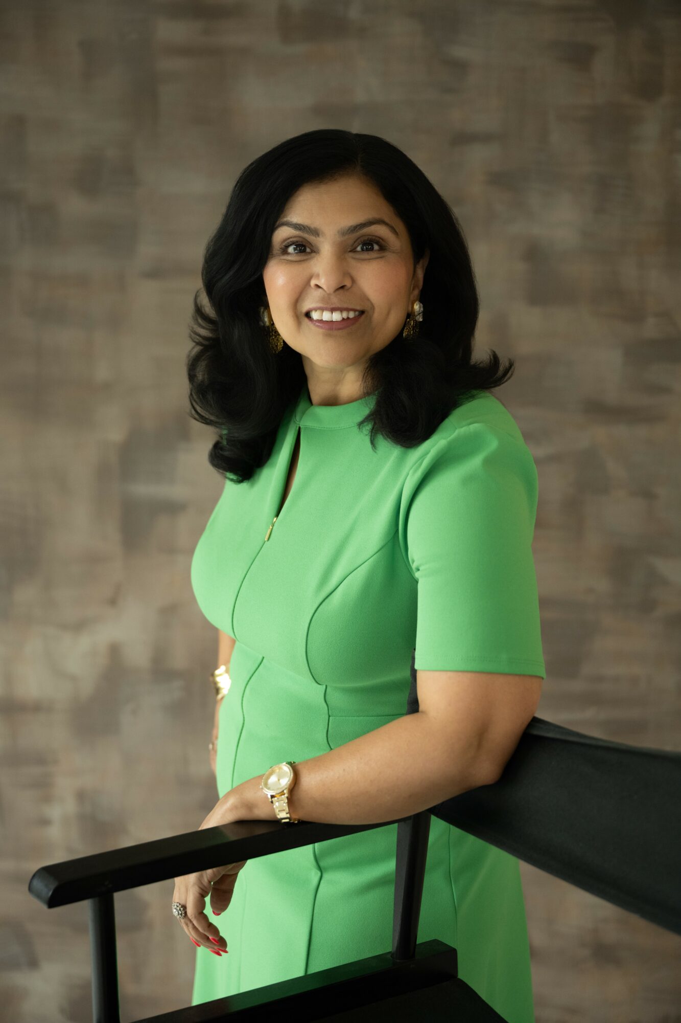 Smiling woman with black hair in a green dress, standing indoors against a textured background.
