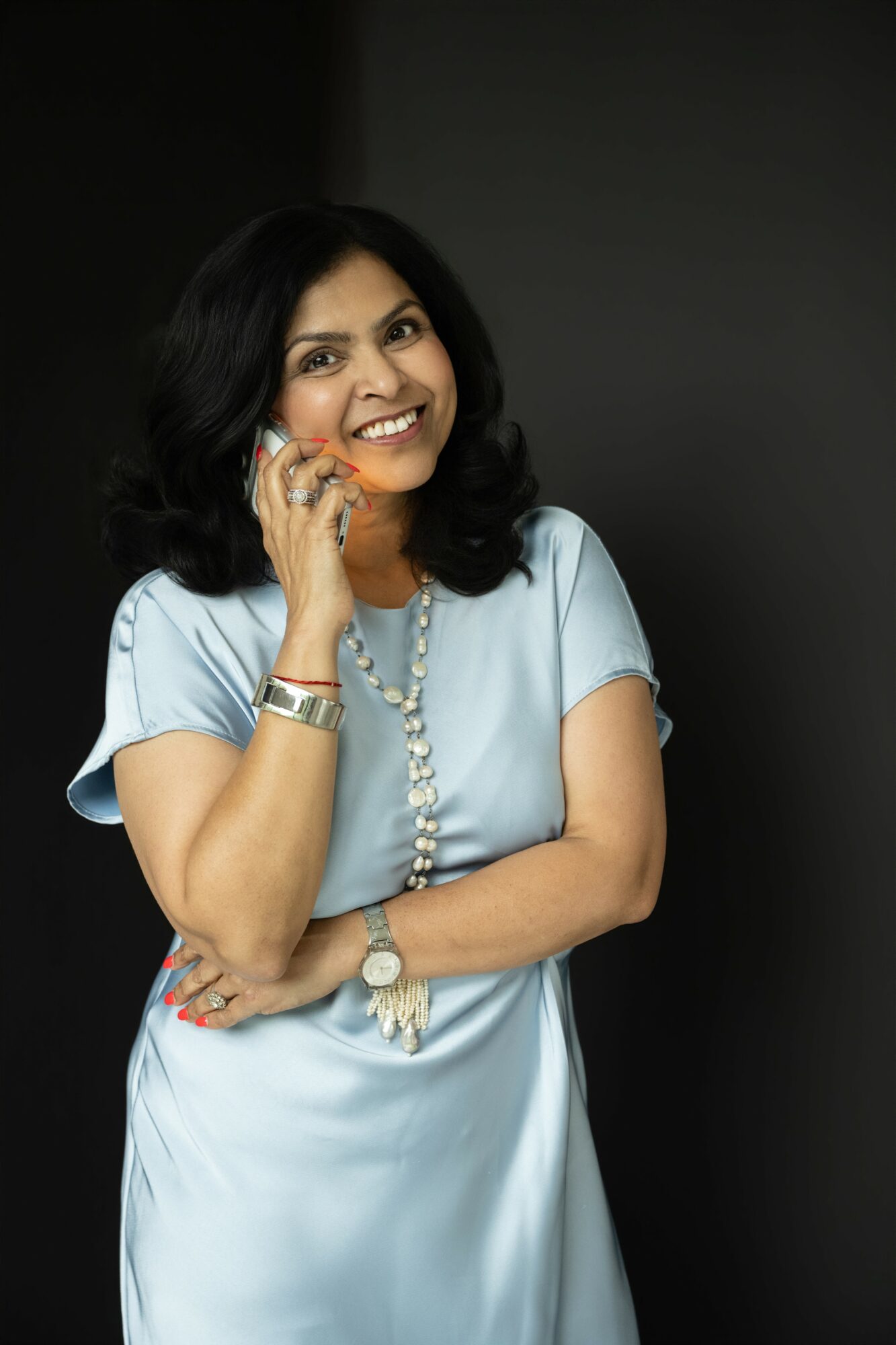 Woman with black hair smiling, wearing a light blue dress, jewelry, and a watch, standing against a dark background.