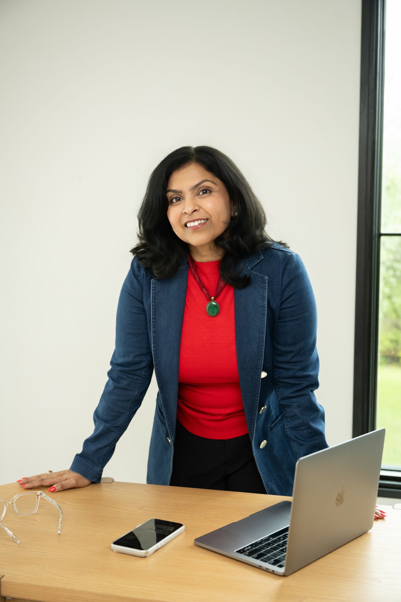 Woman with dark hair in a blue blazer and red top standing at a desk with a laptop and smartphone, smiling.