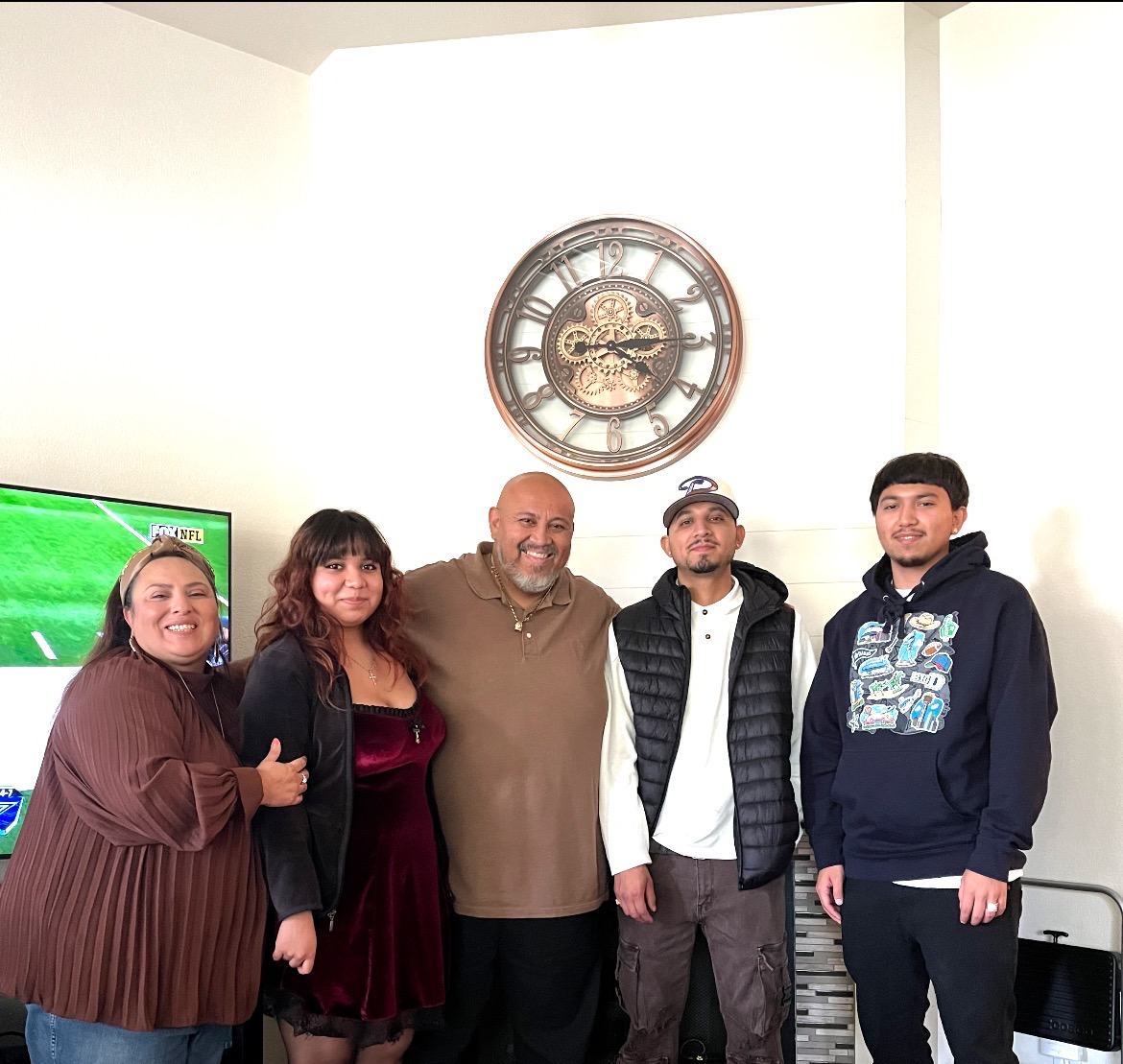 Group of five people standing indoors, smiling, with a large clock on the wall behind them.
