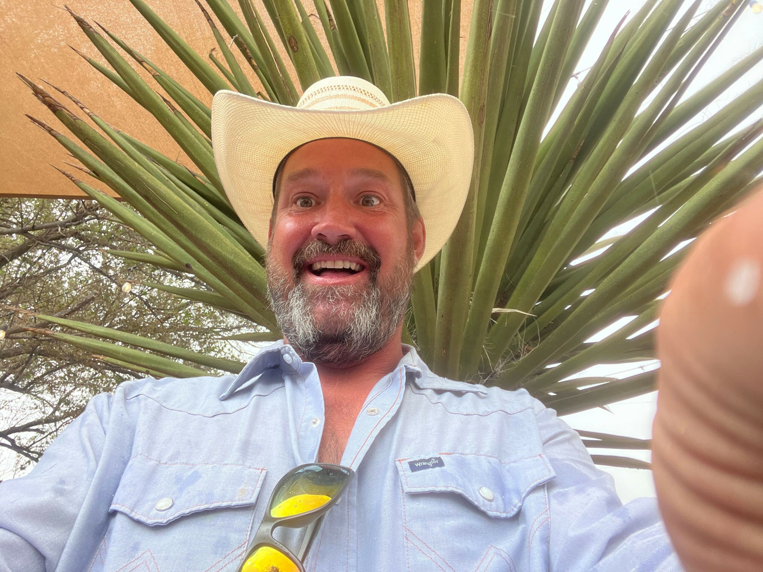 Man wearing a hat and sunglasses, smiling, outdoors with large green plant leaves behind him.