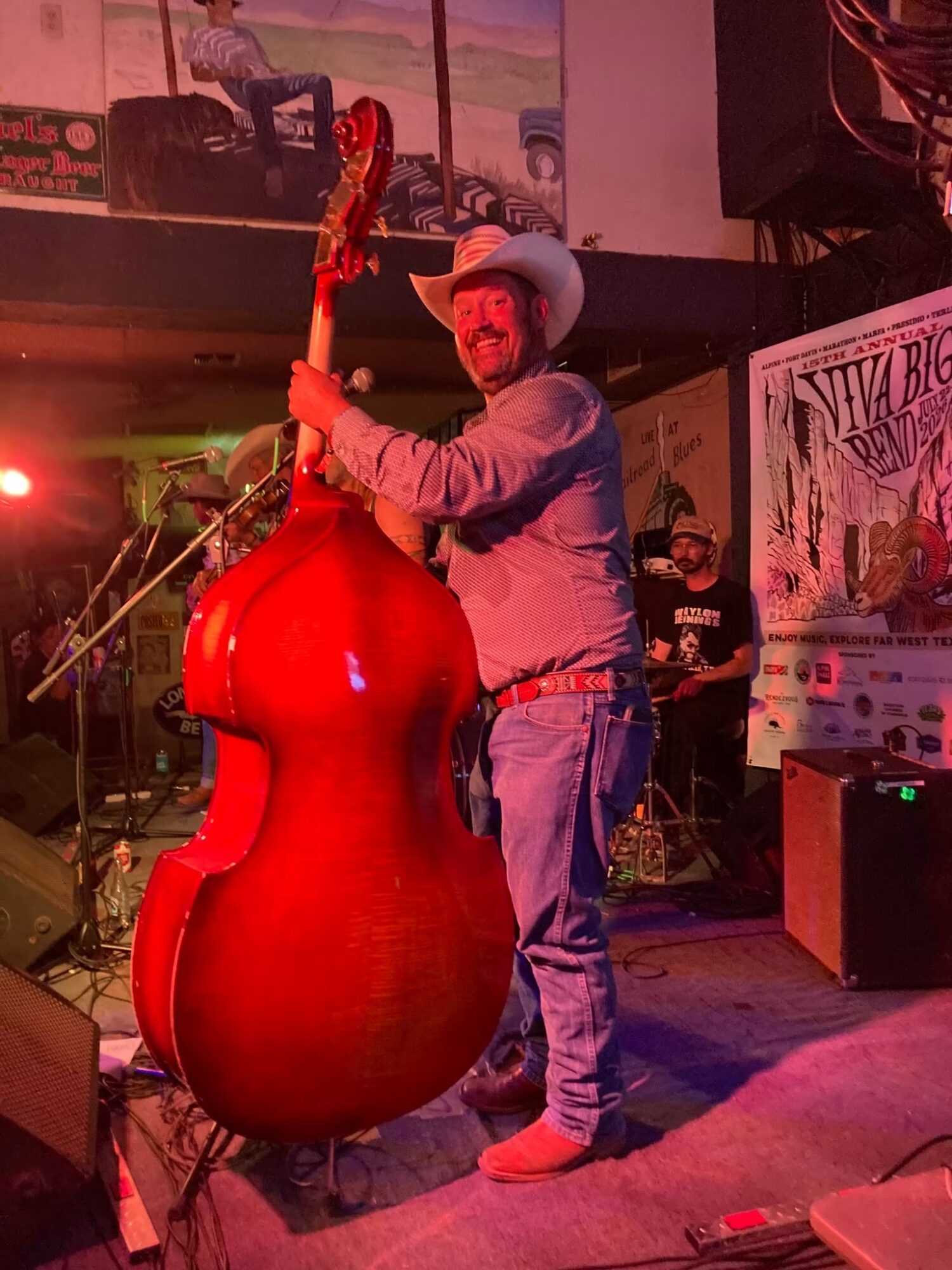 Man in cowboy hat and jeans smiling on stage with a large red guitar-shaped object, microphone, and band members in background.