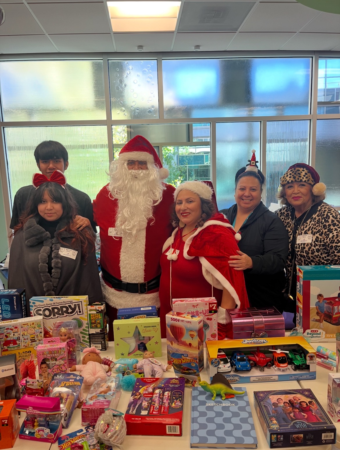 Group of people with Santa Claus and a woman in a red and white costume, standing behind a table with toys, in a room with large windows.