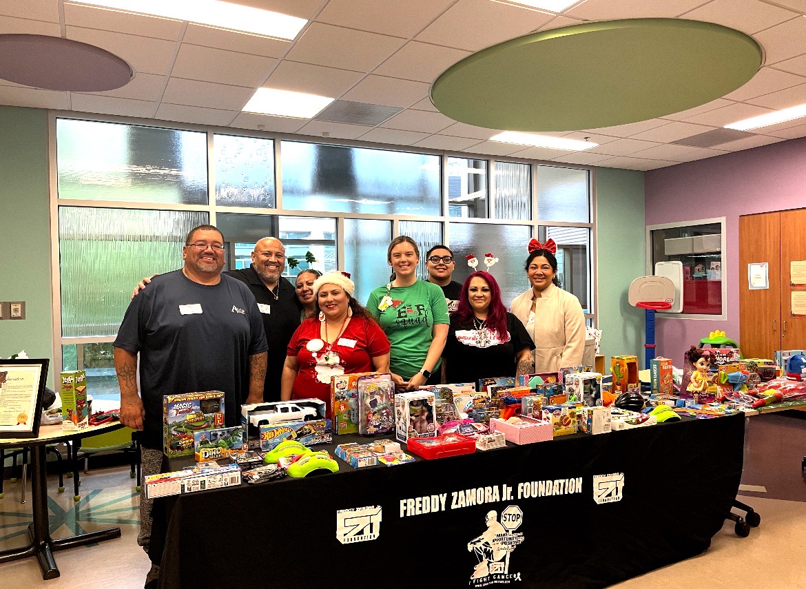 Group of people standing behind a table filled with toys, in a room with large windows and colorful walls.
