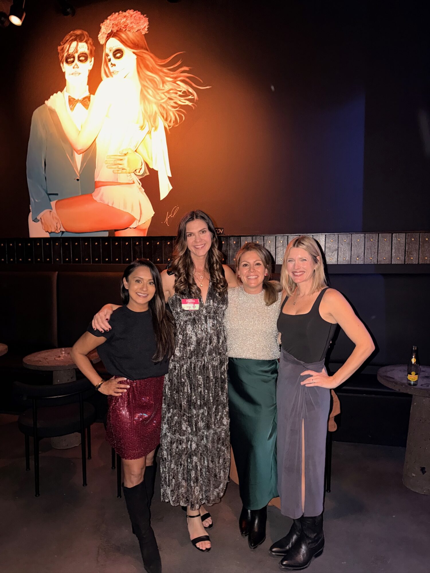 Four women standing together in a dimly lit room, smiling, with a large animated projection of two women on a screen behind them.