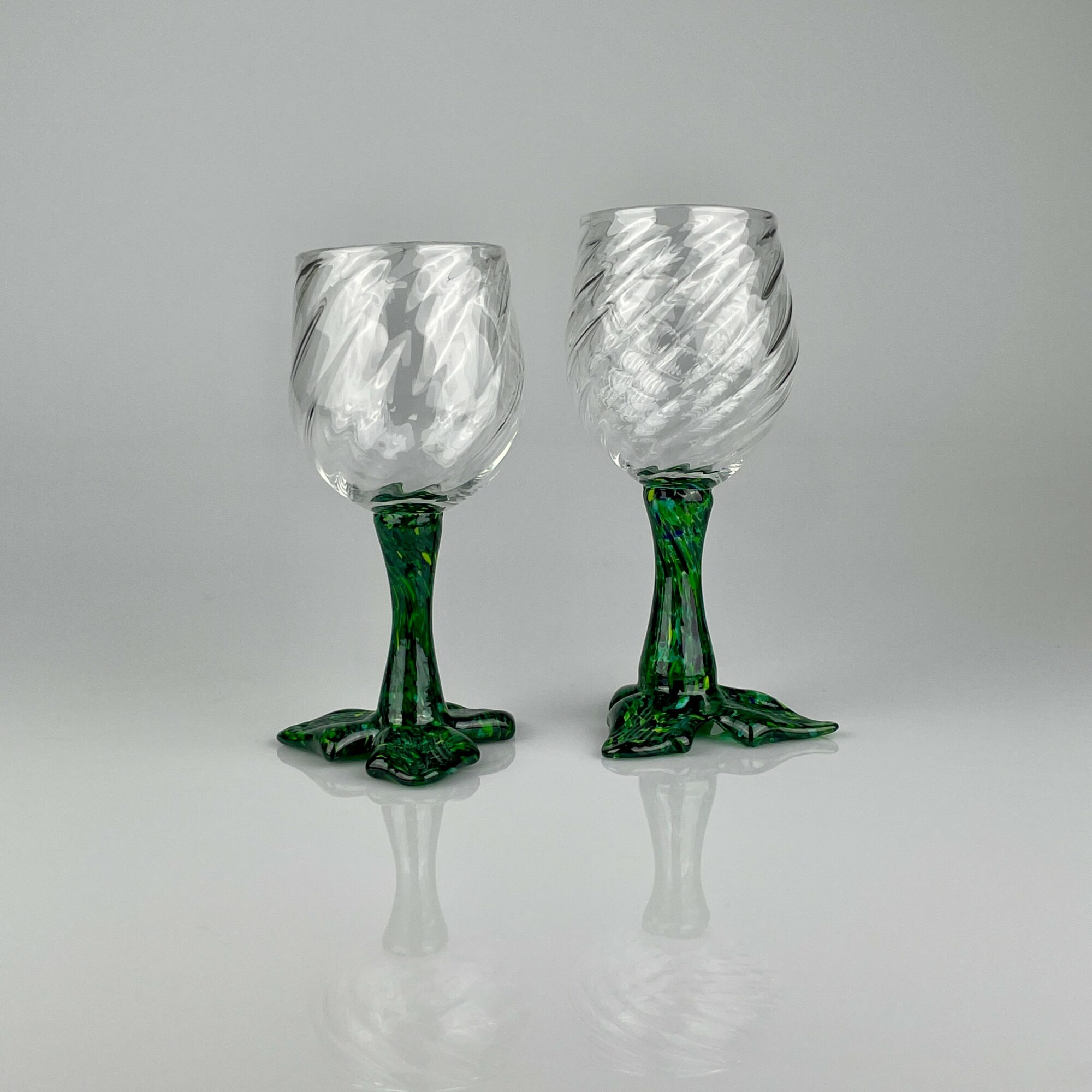Two glass goblets with green bases and clear, textured bowls, placed on a reflective surface against a plain background.