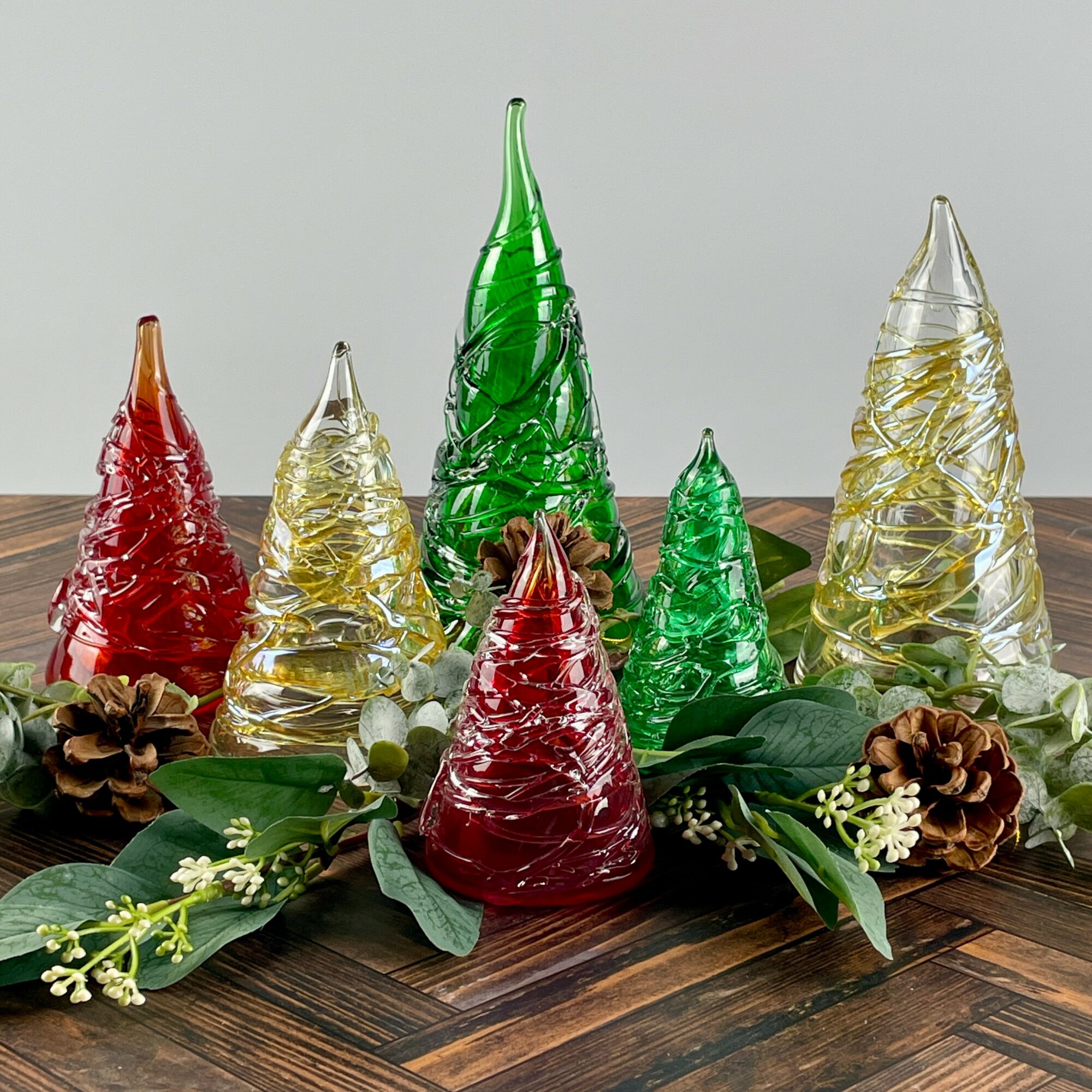 Decorative glass Christmas trees in red, green, and clear colors arranged with pinecones and greenery on a wooden surface.