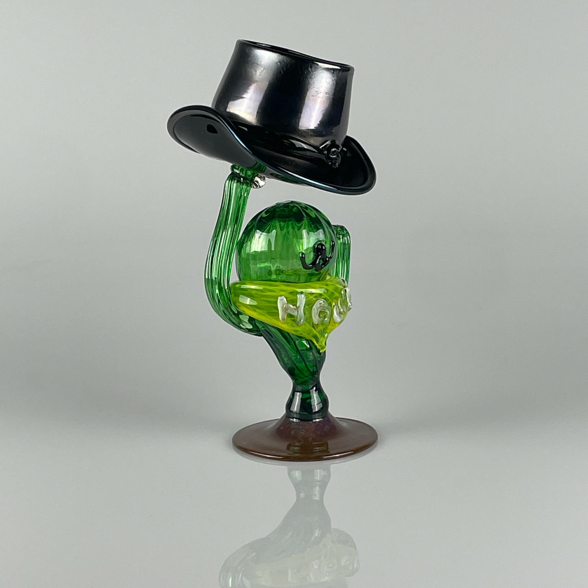 Glass sculpture of a green figure with a black top hat, standing on a round base, holding the hat with one hand.