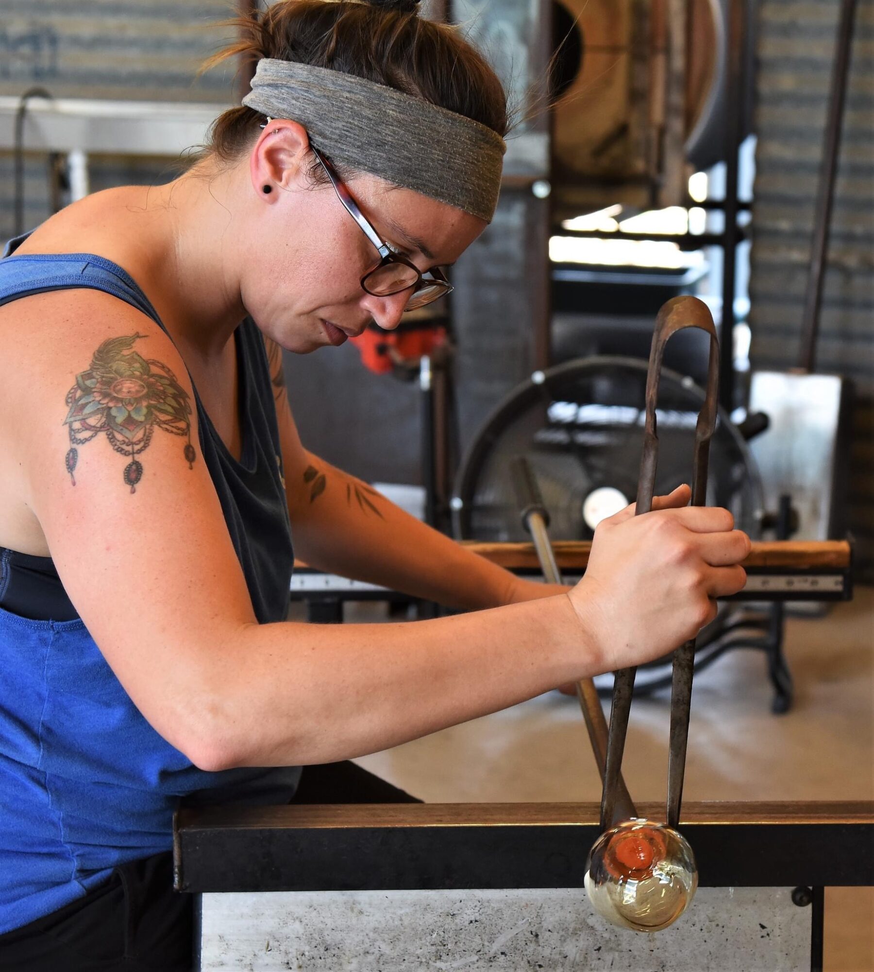 Woman with glasses and headband working on a glassblowing project, holding tools near a glass object.