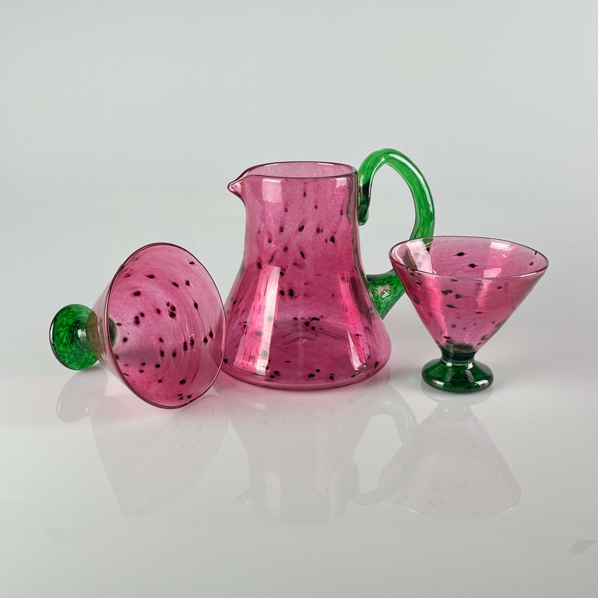 Pink glass pitcher, bowl, and cup with green accents on a white surface, reflected below. Clear and colorful design.