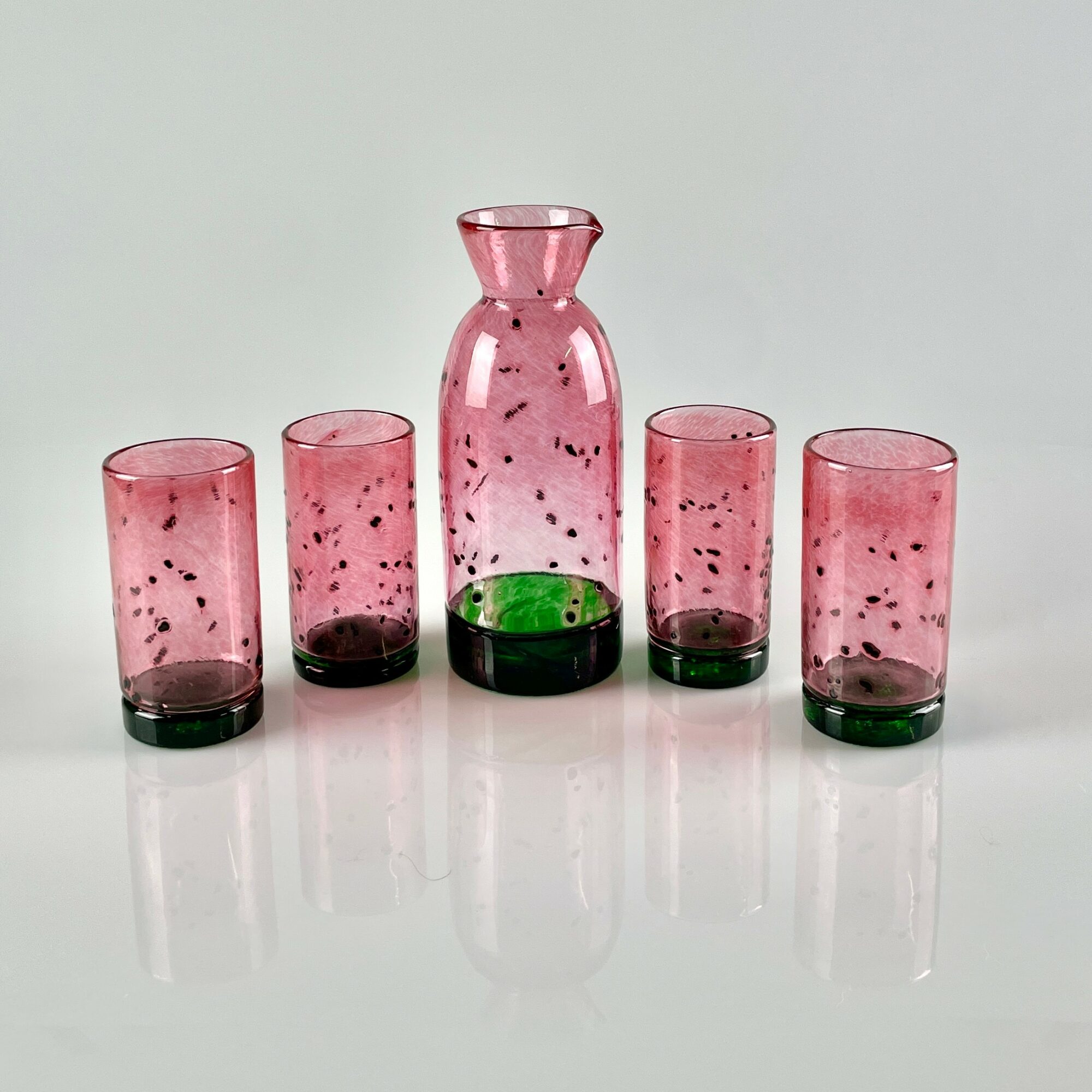Pink glass pitcher with green base surrounded by four matching pink glasses, all with black speckles, on a reflective surface.
