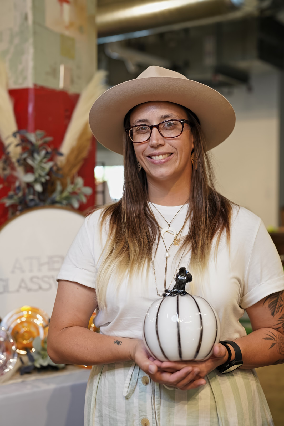 Smiling woman wearing glasses and a wide-brimmed hat holding a decorative pumpkin, standing indoors with plants and decorations in background.