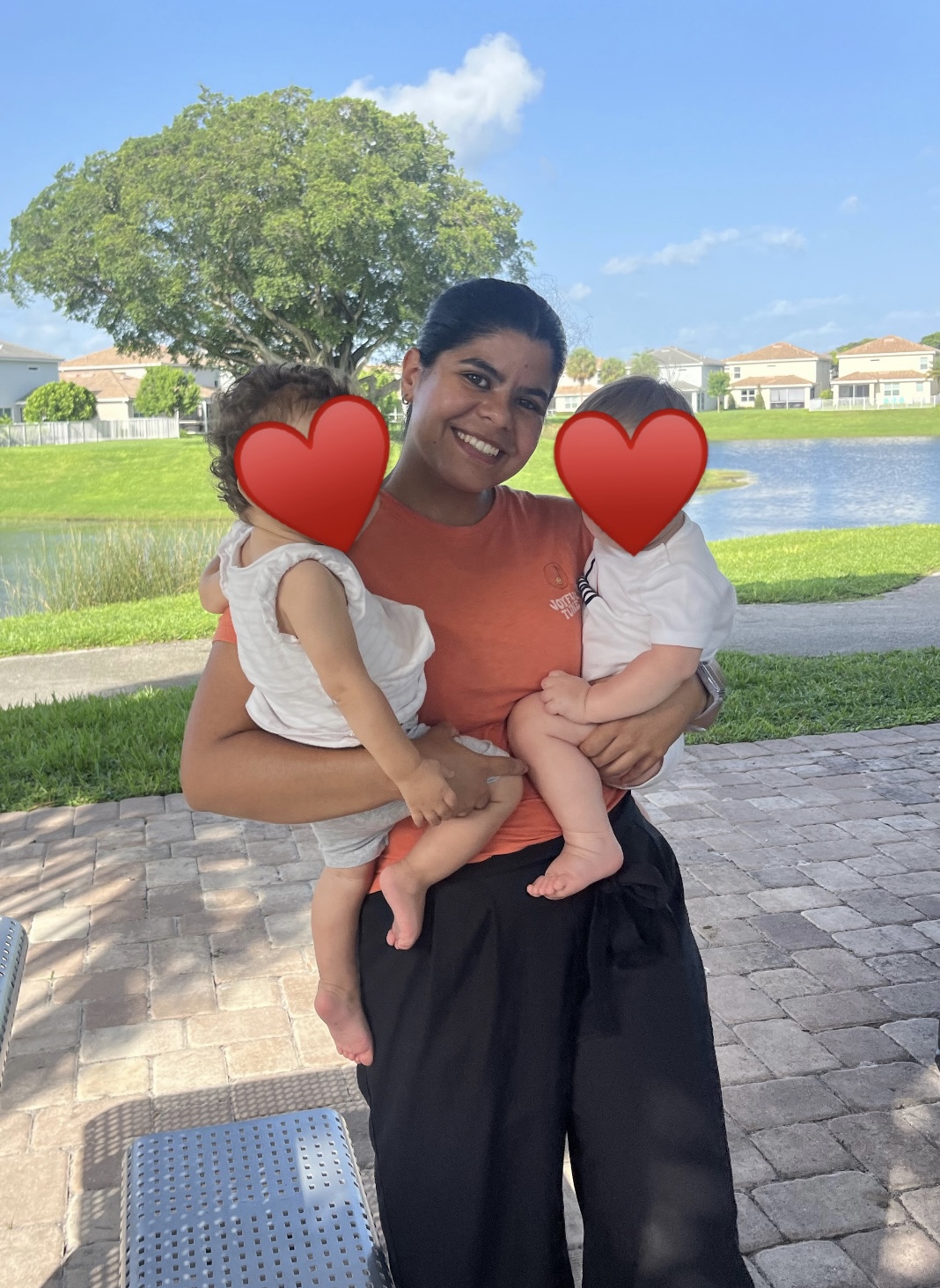 Woman holding two children outdoors near a lake with trees and houses in background.