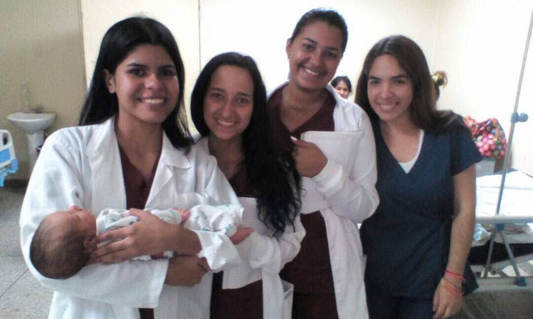 Four women, one holding a newborn, smiling in a hospital room, with medical equipment in the background.