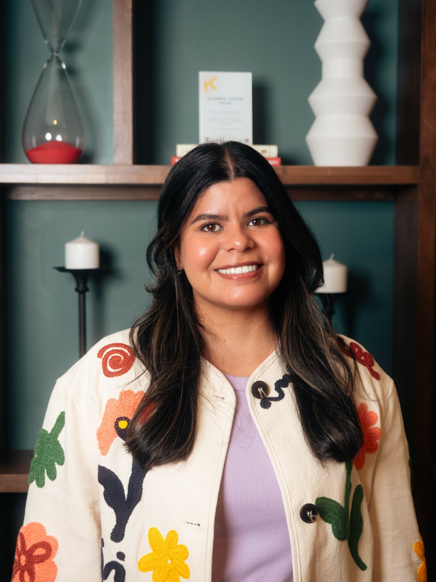 Woman with long dark hair smiling, standing indoors with decorative vases and candles in the background.