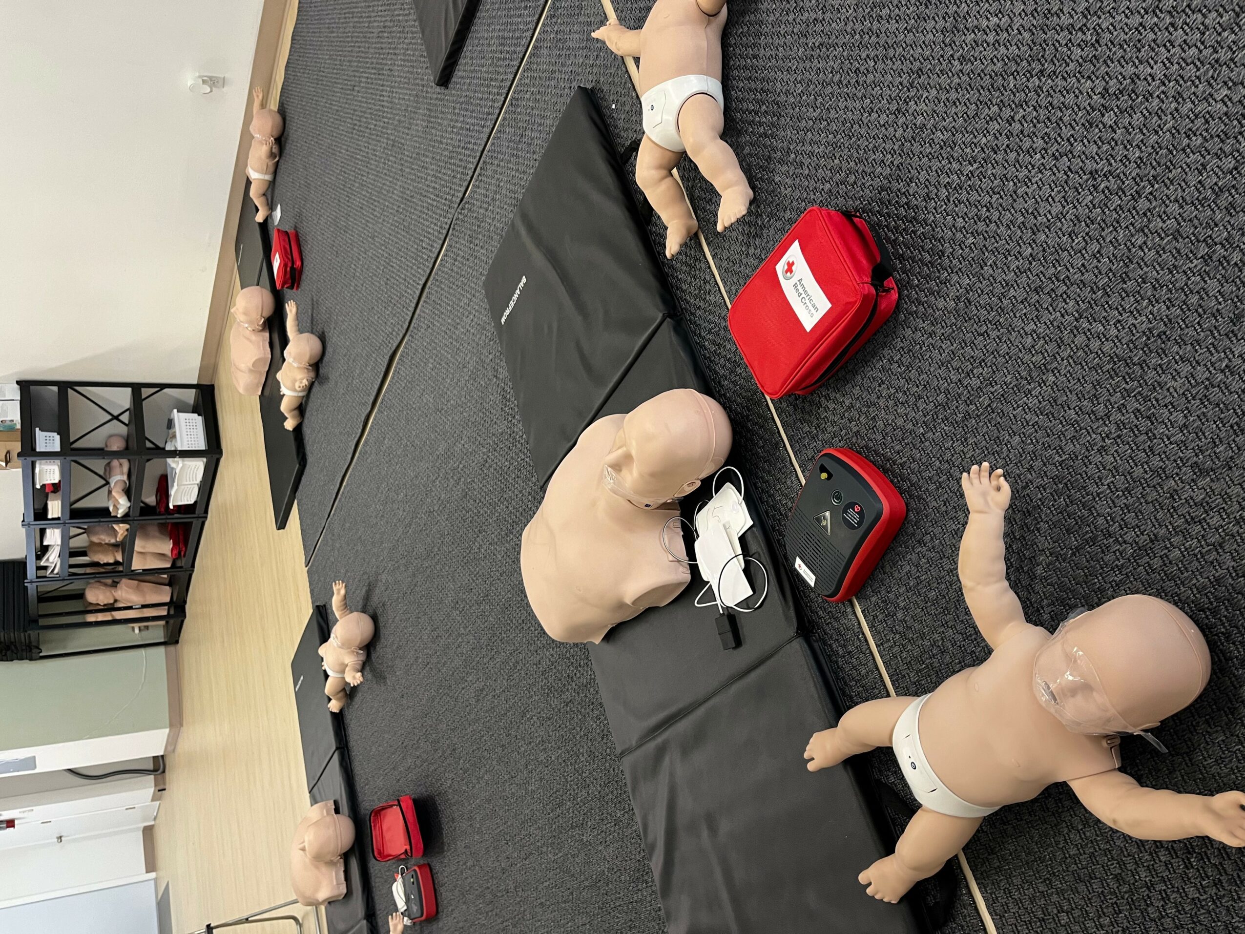 Multiple baby dolls and a red first aid kit on a black mat in a room with a bookshelf and wooden floor.