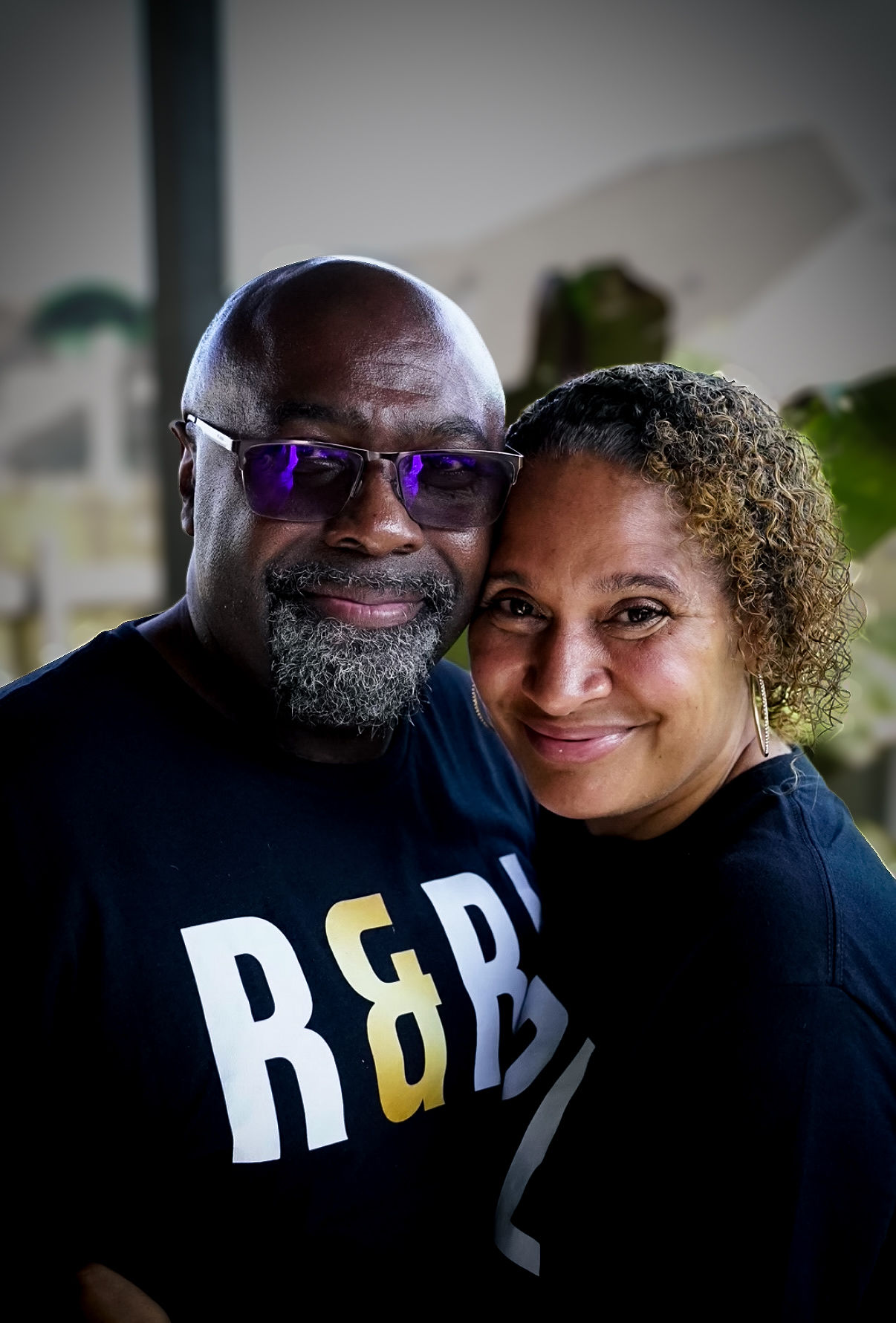 A smiling man and woman close together, indoors, with blurred background, both facing camera.