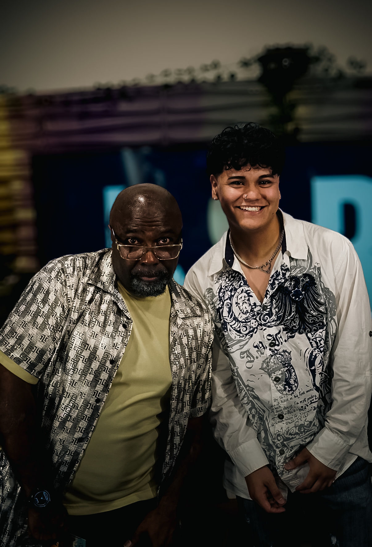 Two men standing together, smiling, indoors with a blurred background. One man has glasses and a patterned shirt, the other has curly hair and a white shirt with a design.