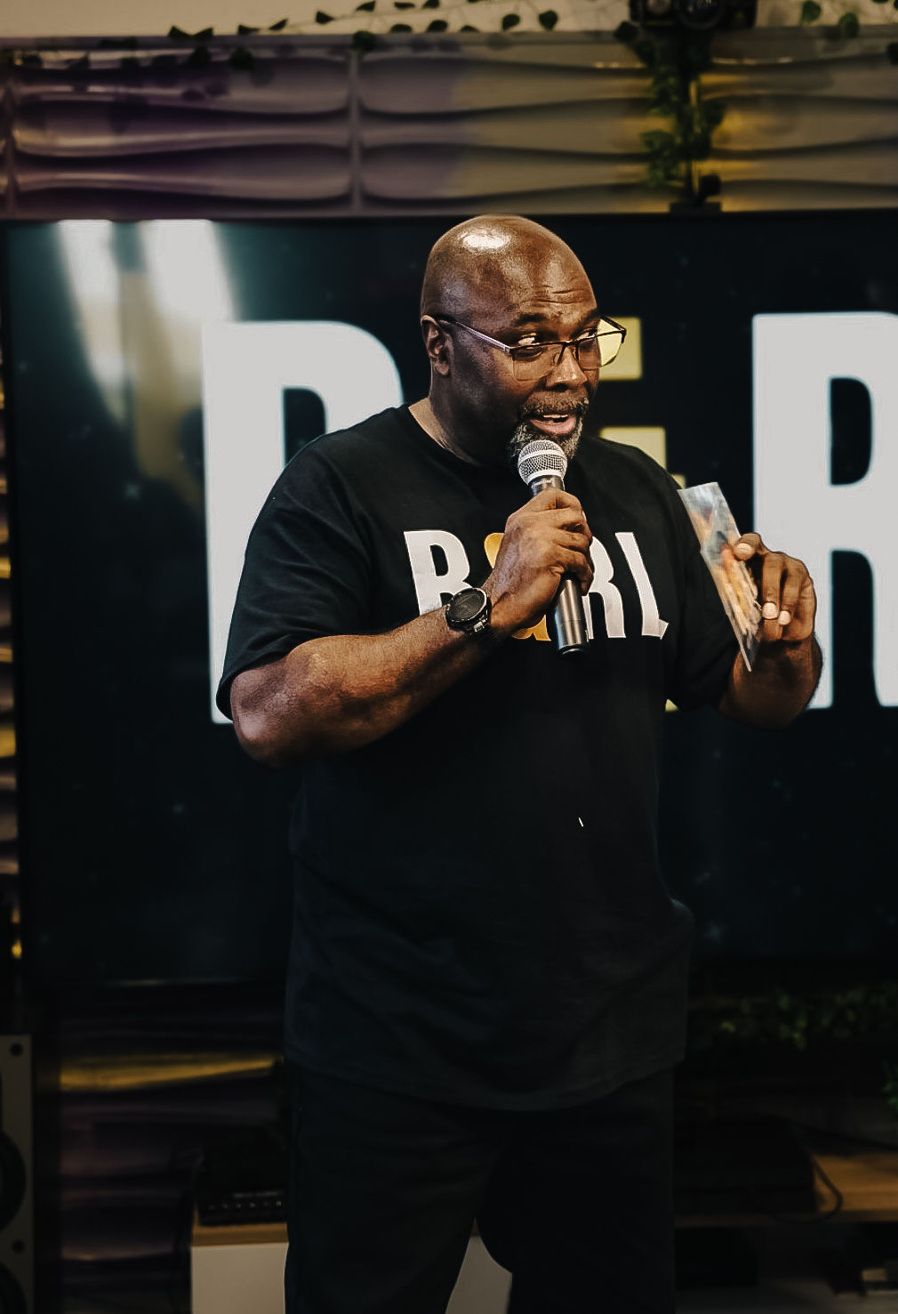 Man speaking into microphone on stage, wearing glasses and a black T-shirt, with large letters in the background.