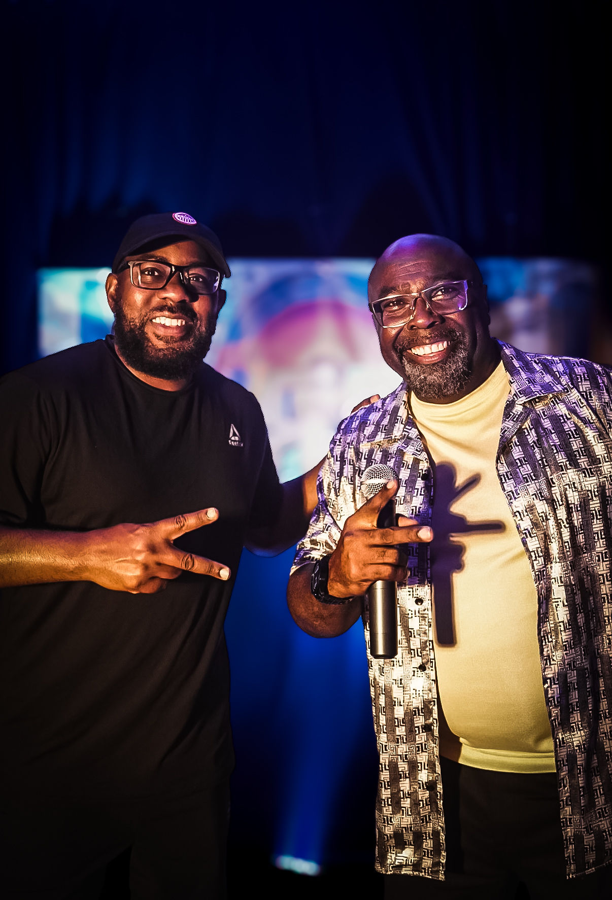Two smiling men with glasses, one in a black shirt and the other in a patterned shirt, holding microphones on stage.