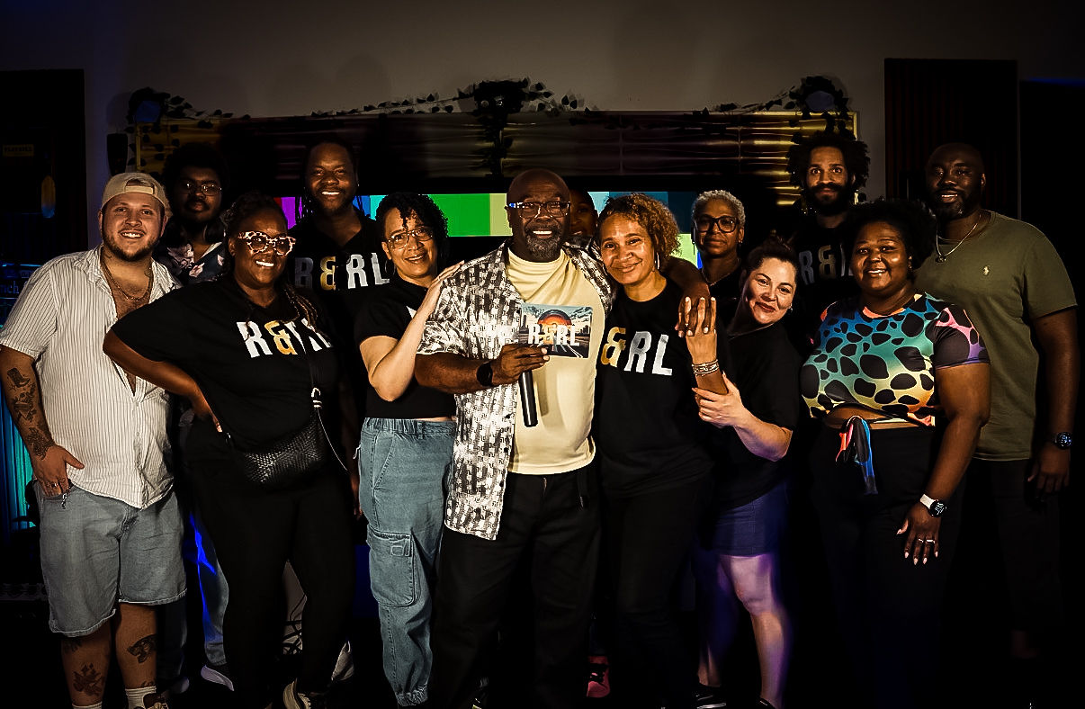 Group of diverse people smiling and posing together indoors, some making gestures, with a dark background.
