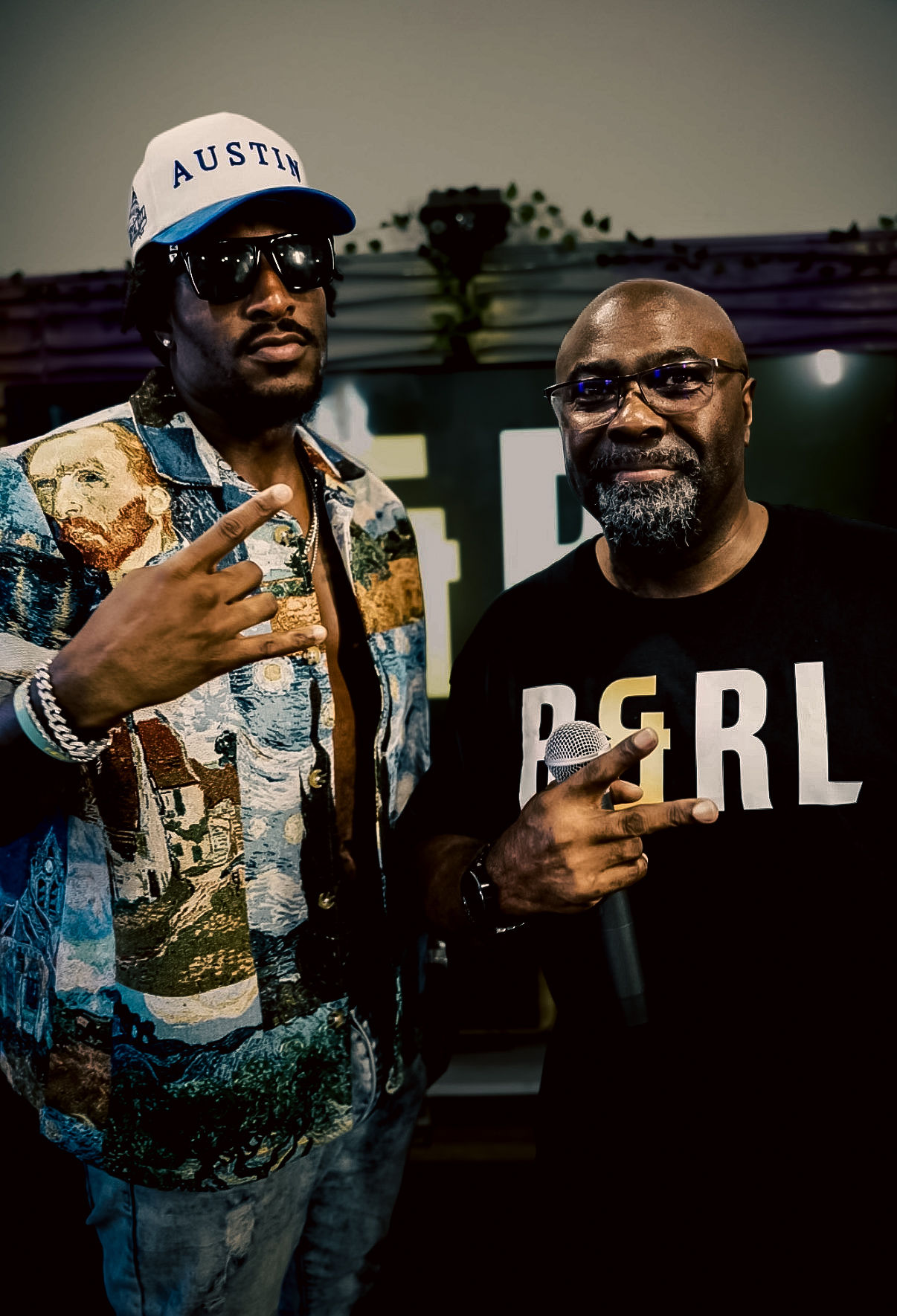 Two men pose together, one wearing sunglasses, a cap, and a patterned jacket, the other in a black shirt with 'BRL' text, both making hand gestures.