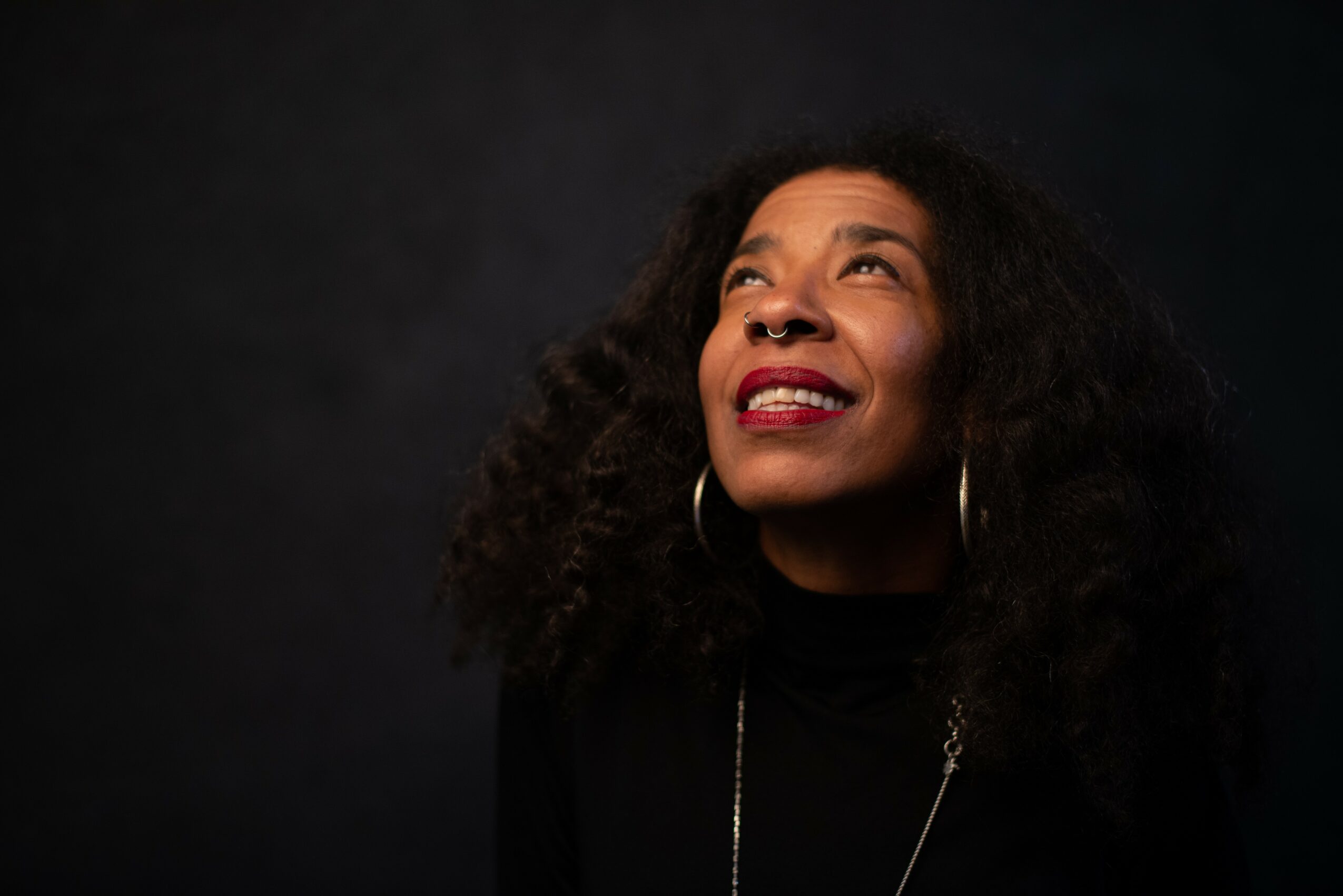Woman with curly hair smiling and looking upward, wearing hoop earrings and a black top, dark background.