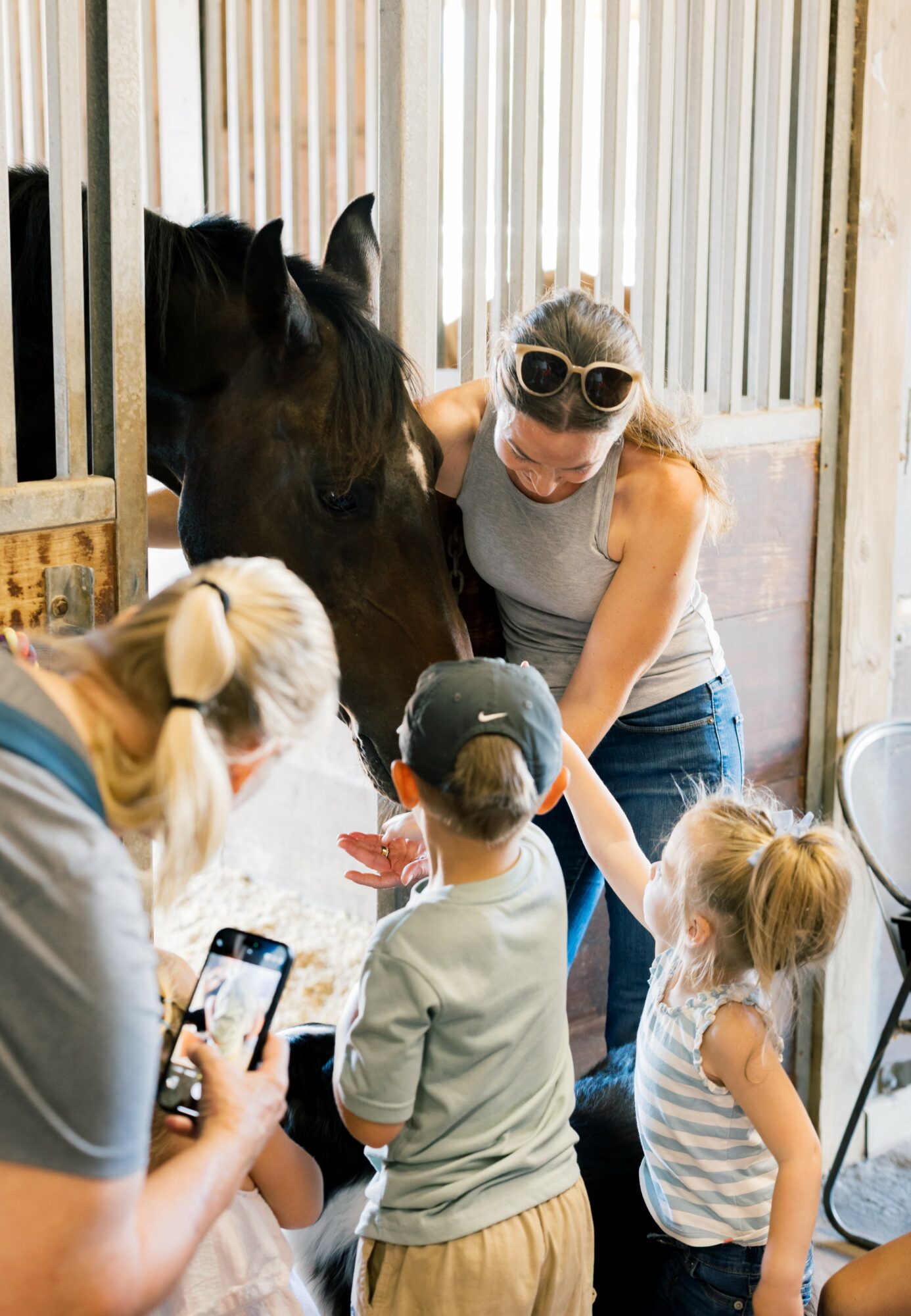 Woman with sunglasses interacts with a horse while children and a person take photos nearby in a barn.
