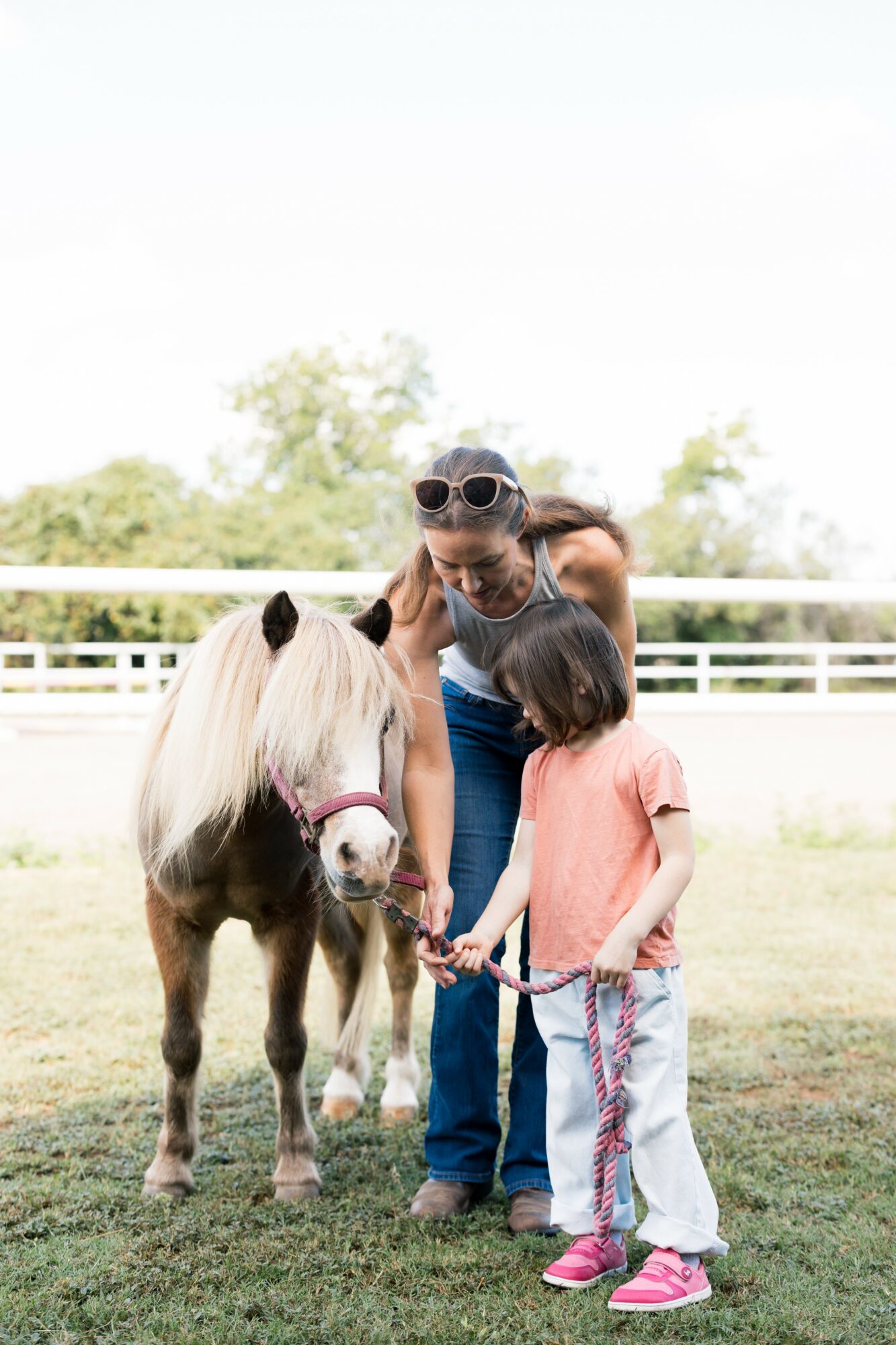 Two children and a woman with sunglasses stand outdoors with a small pony, holding its lead rope, on a grassy field.