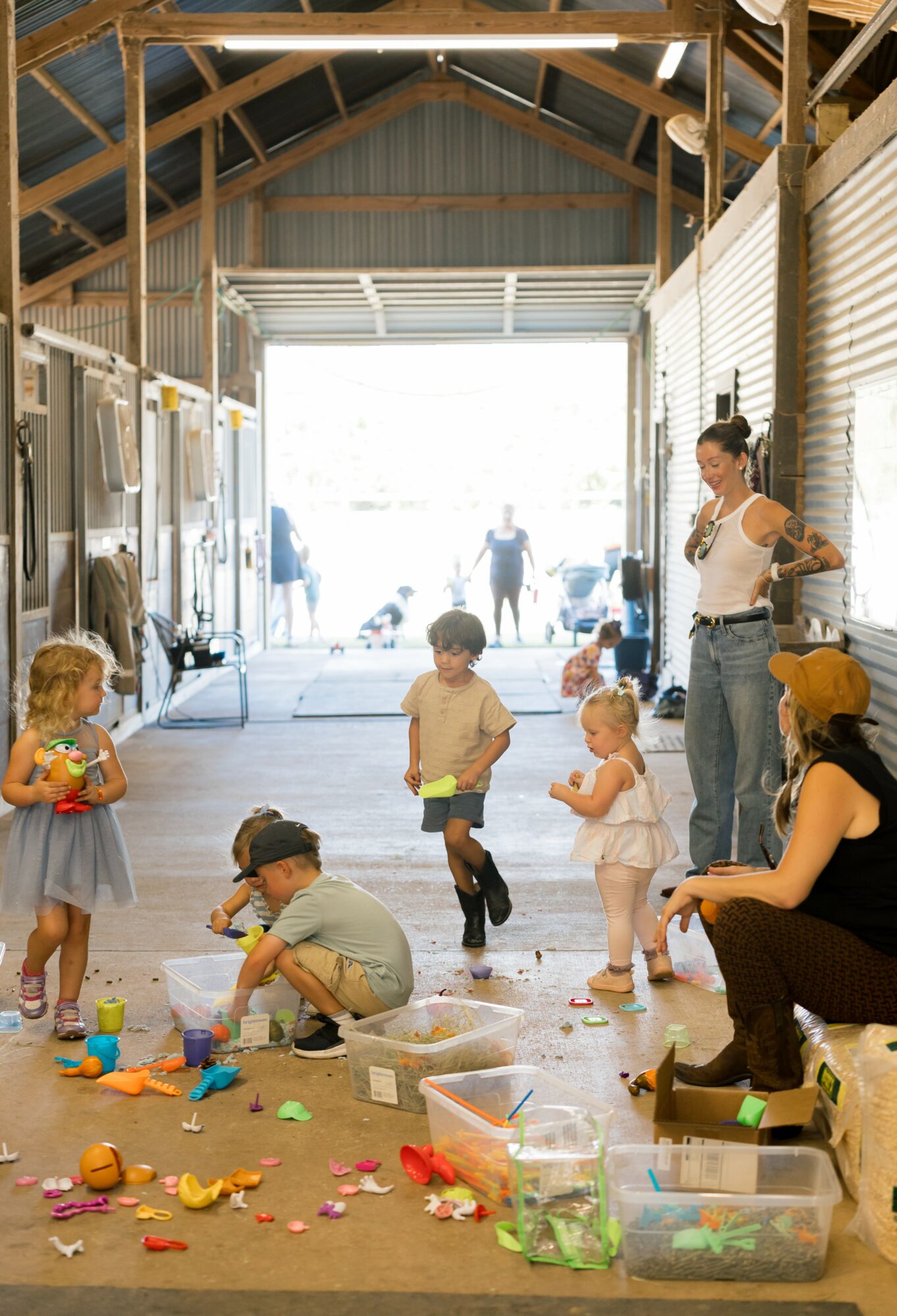 Children playing with toys inside a barn-like structure, with a woman supervising, and people outside visible through open doors.