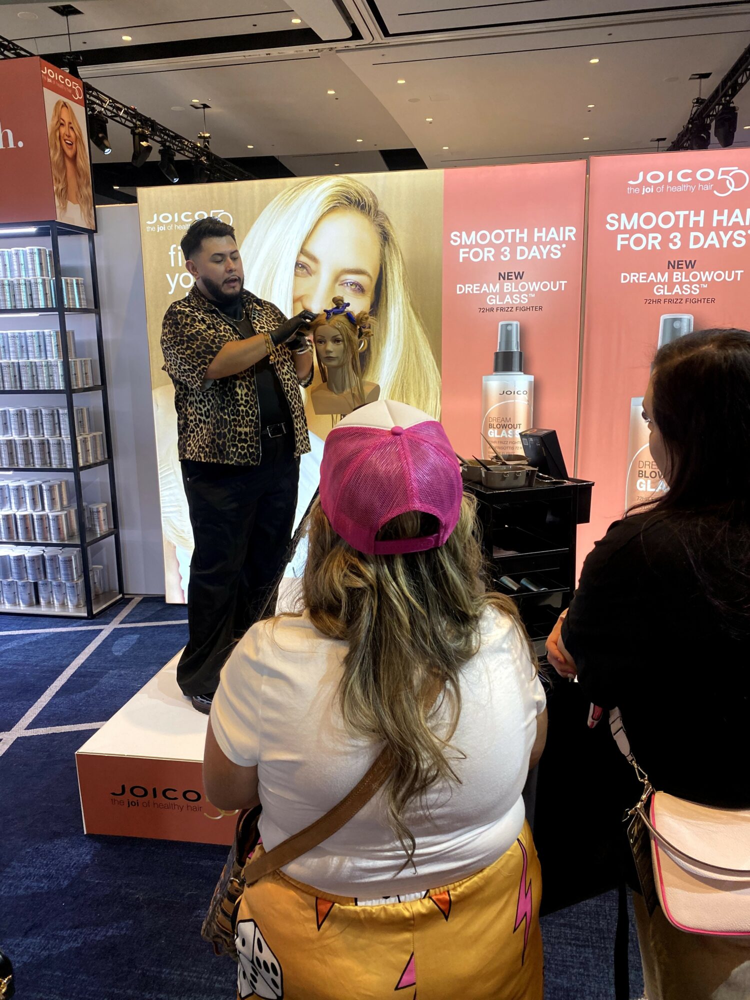 Man presenting hair product to audience in store display with large woman's face and product image behind him.