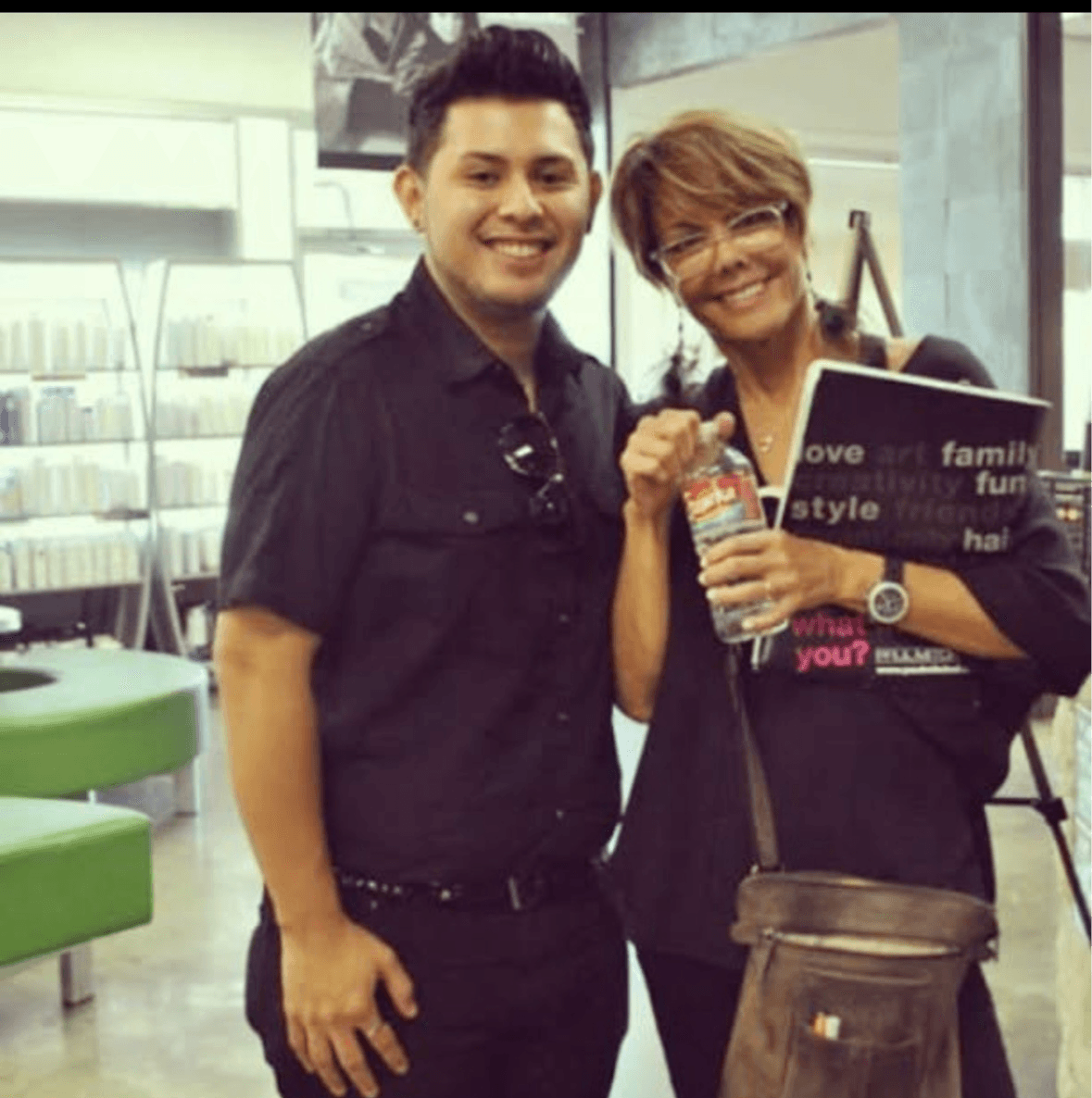 Two people smiling, one holding a water bottle, in a store with shelves and green seating in the background.